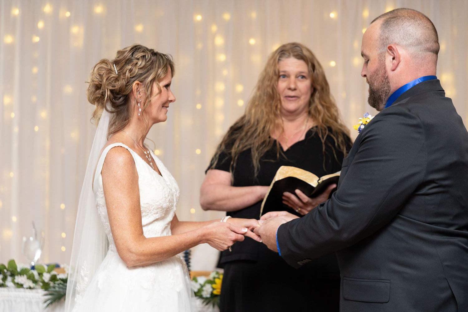 Bride and groom exchanging vows at wedding ceremony with officiant holding bible, fairy lights backdrop.