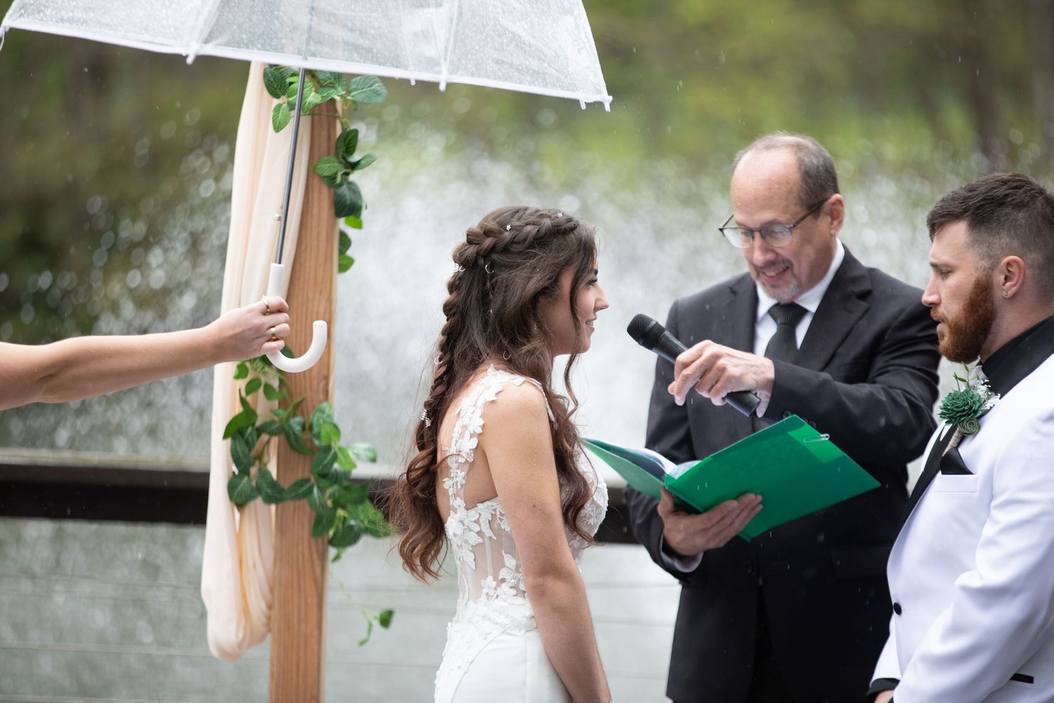 Bride and groom exchange vows with officiant holding green book during outdoor rainy wedding ceremony.