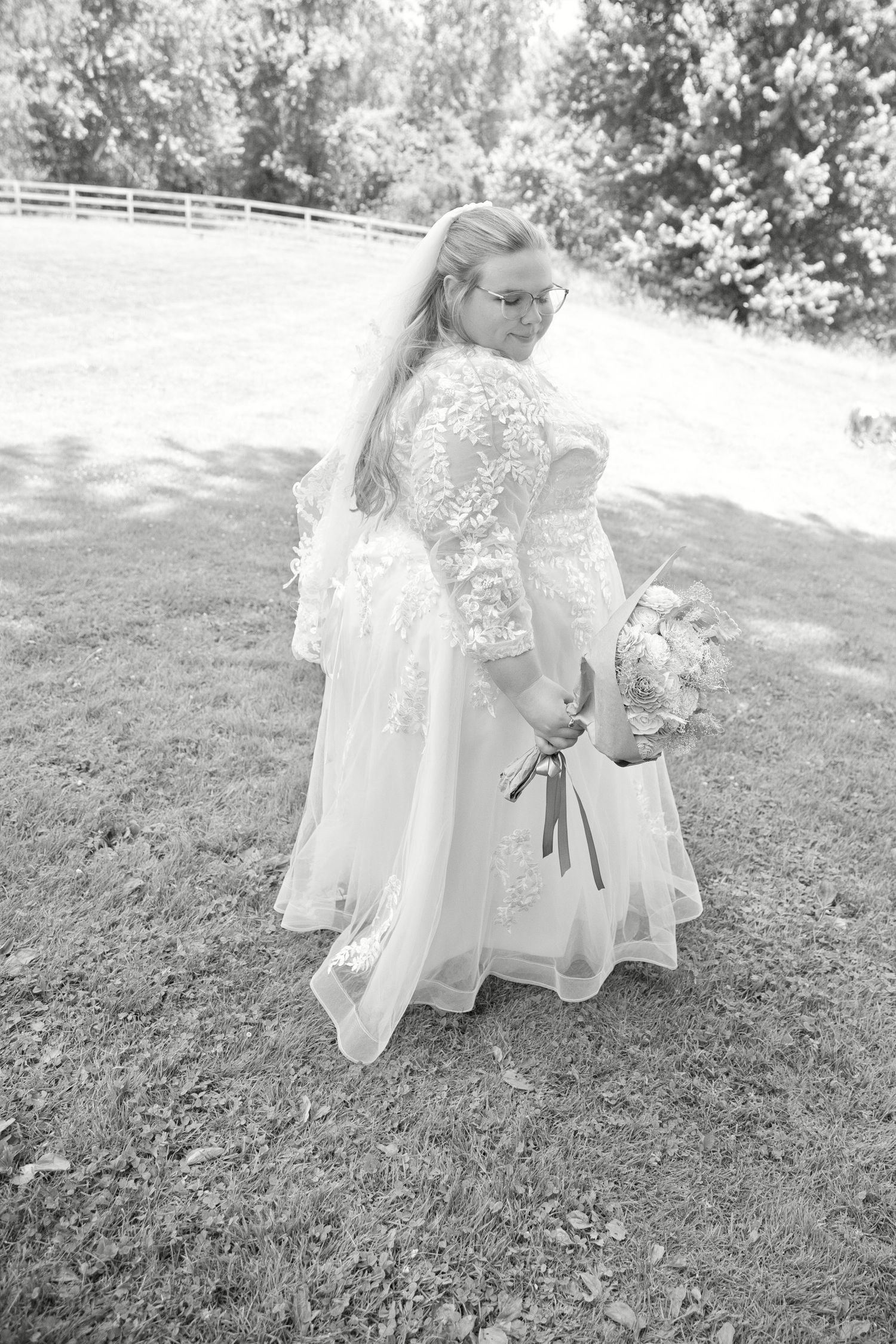 Black and white photo of a bride in lace gown holding bouquet, standing on grass with trees and fence backdrop.