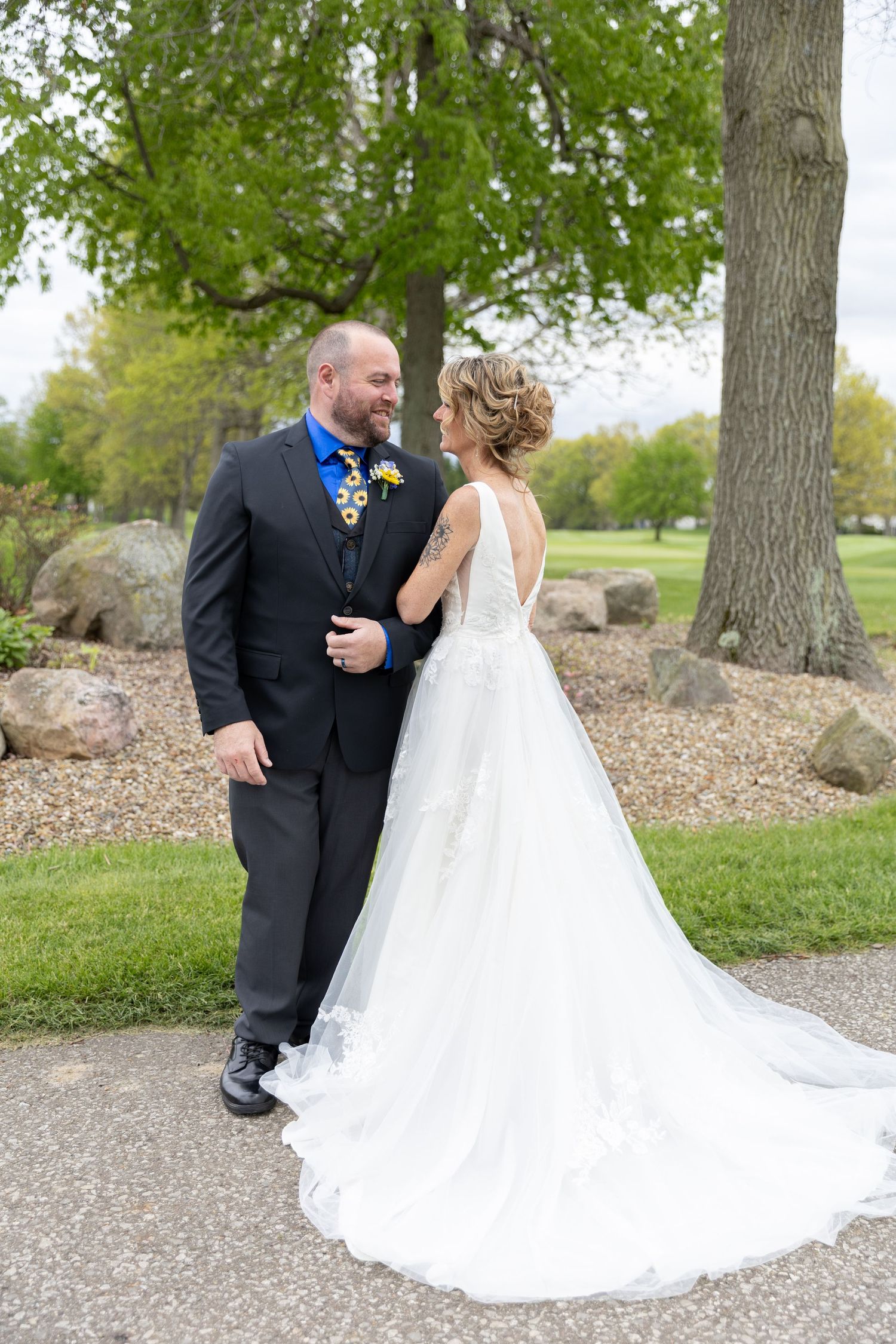 Bride and groom share a romantic moment outdoors, she in a white lace gown, he in a dark suit with blue tie.