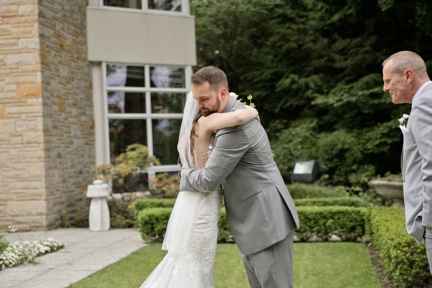 Bride and groom share an emotional first look hug outdoors at a garden wedding venue, with a witness standing nearby.
