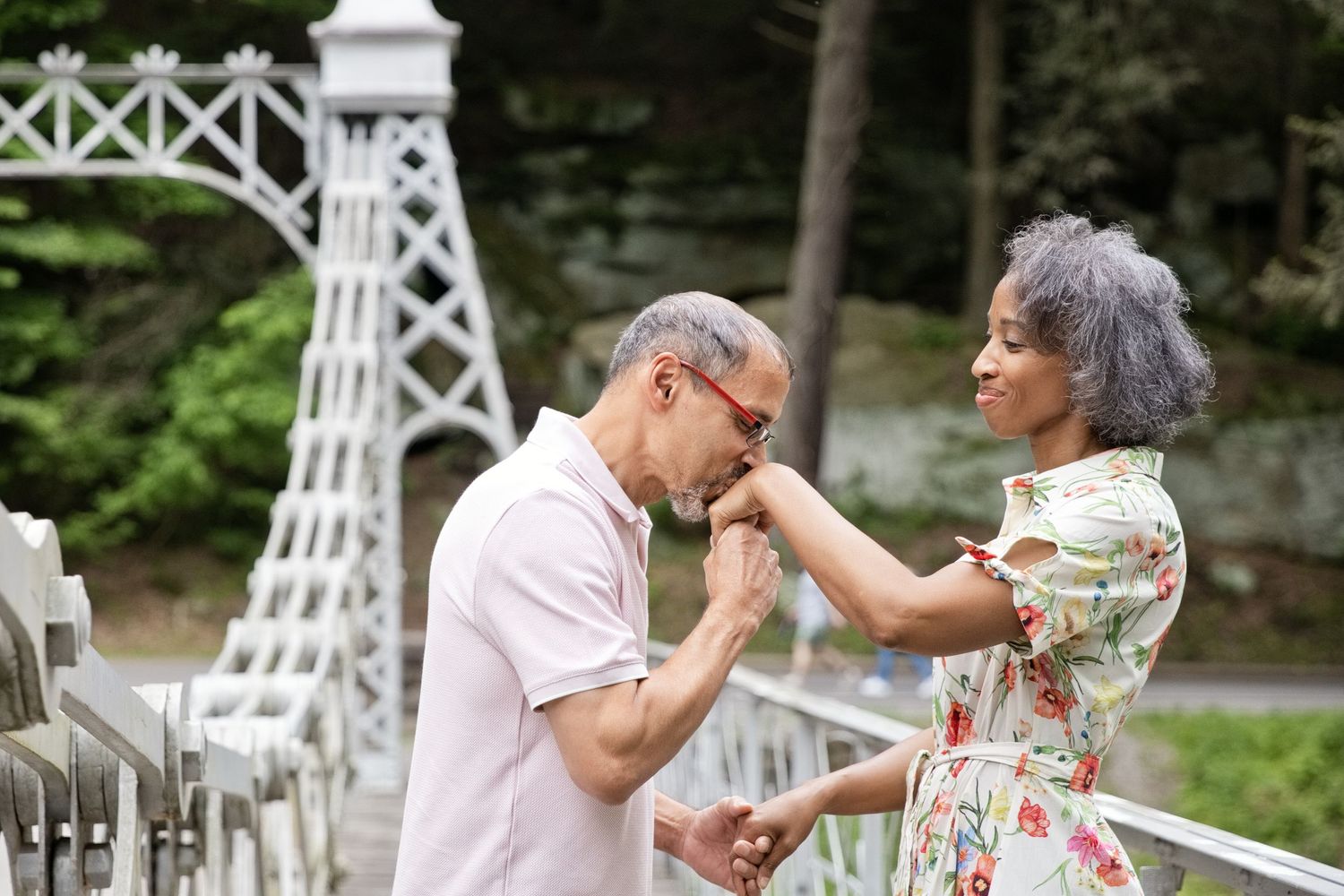 Elderly couple sharing a romantic moment, man kissing woman's hand near white bridge in lush park.
