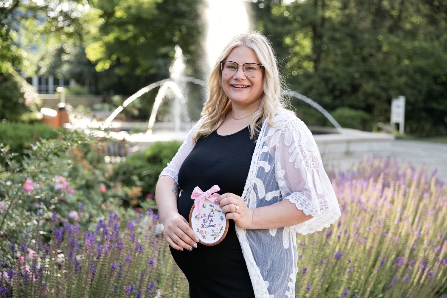 Pregnant woman in black dress and lace cardigan holding a baby shower cookie in a garden with lavender and fountain.
