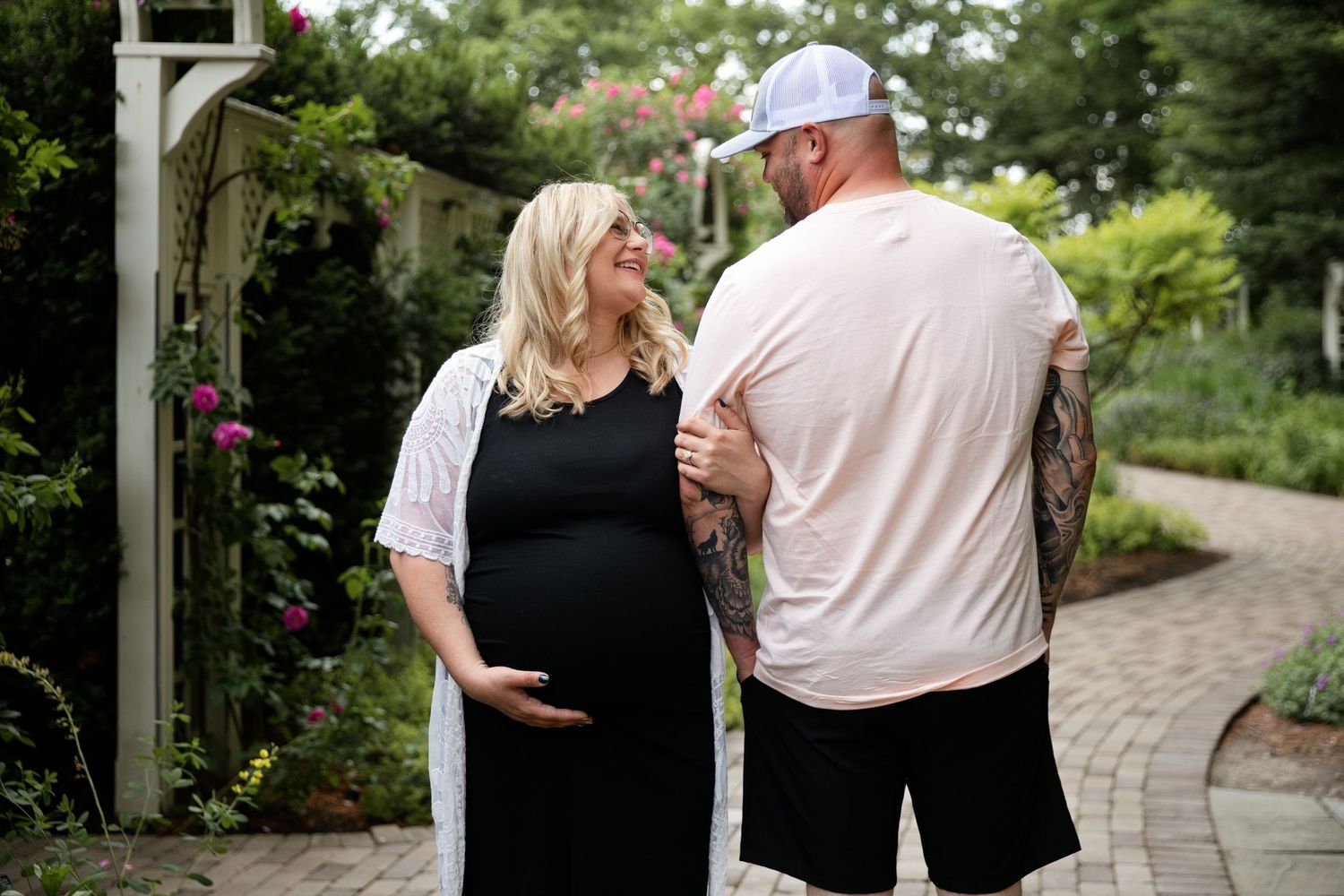 Pregnant couple smiling at each other in a lush garden, man with tattoos in white tee, woman in black maternity dress.