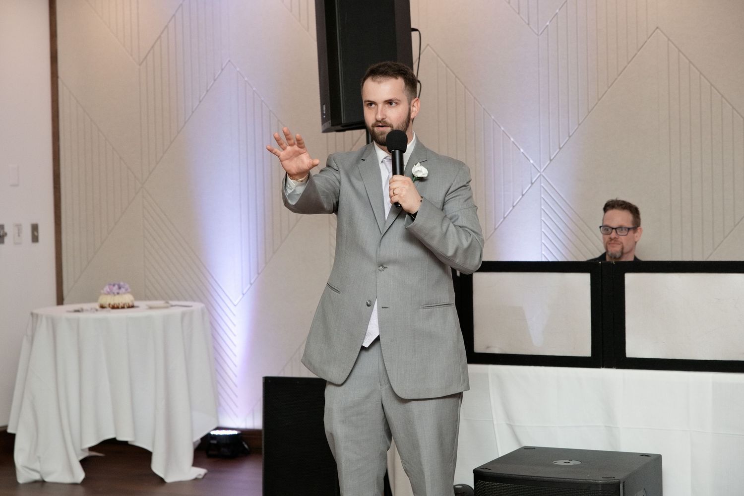 Man in gray suit speaking into microphone at formal event with DJ booth in background.