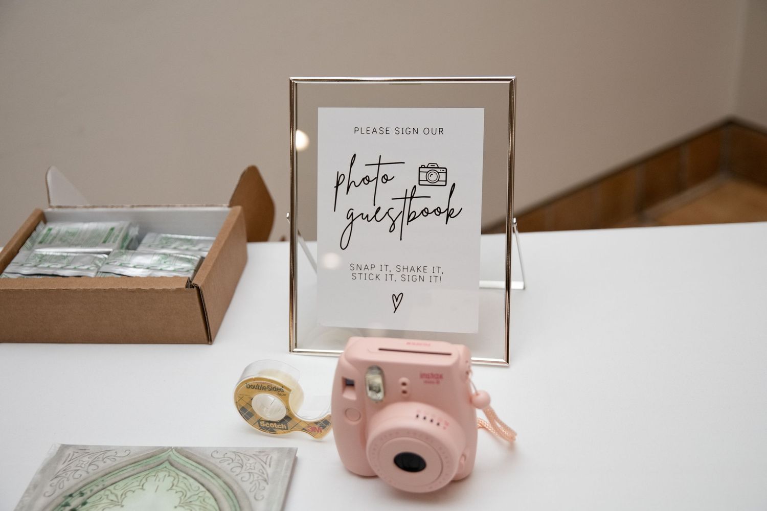 Pink Instax mini camera beside a photo guestbook sign on a white table at a wedding reception.