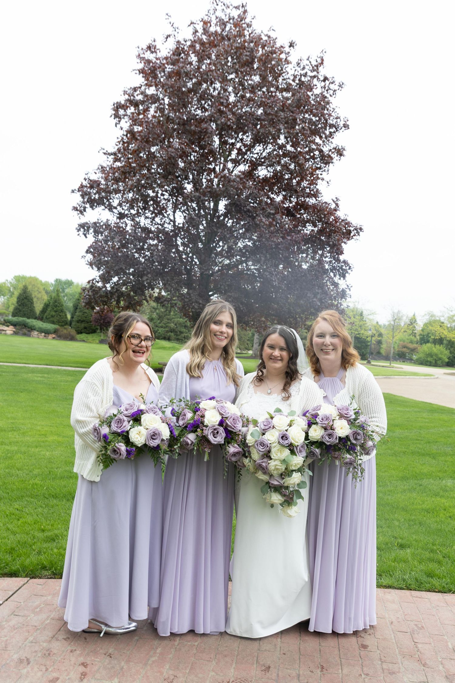 Bride in white gown with three bridesmaids in lavender dresses holding purple and white bouquets outdoors.