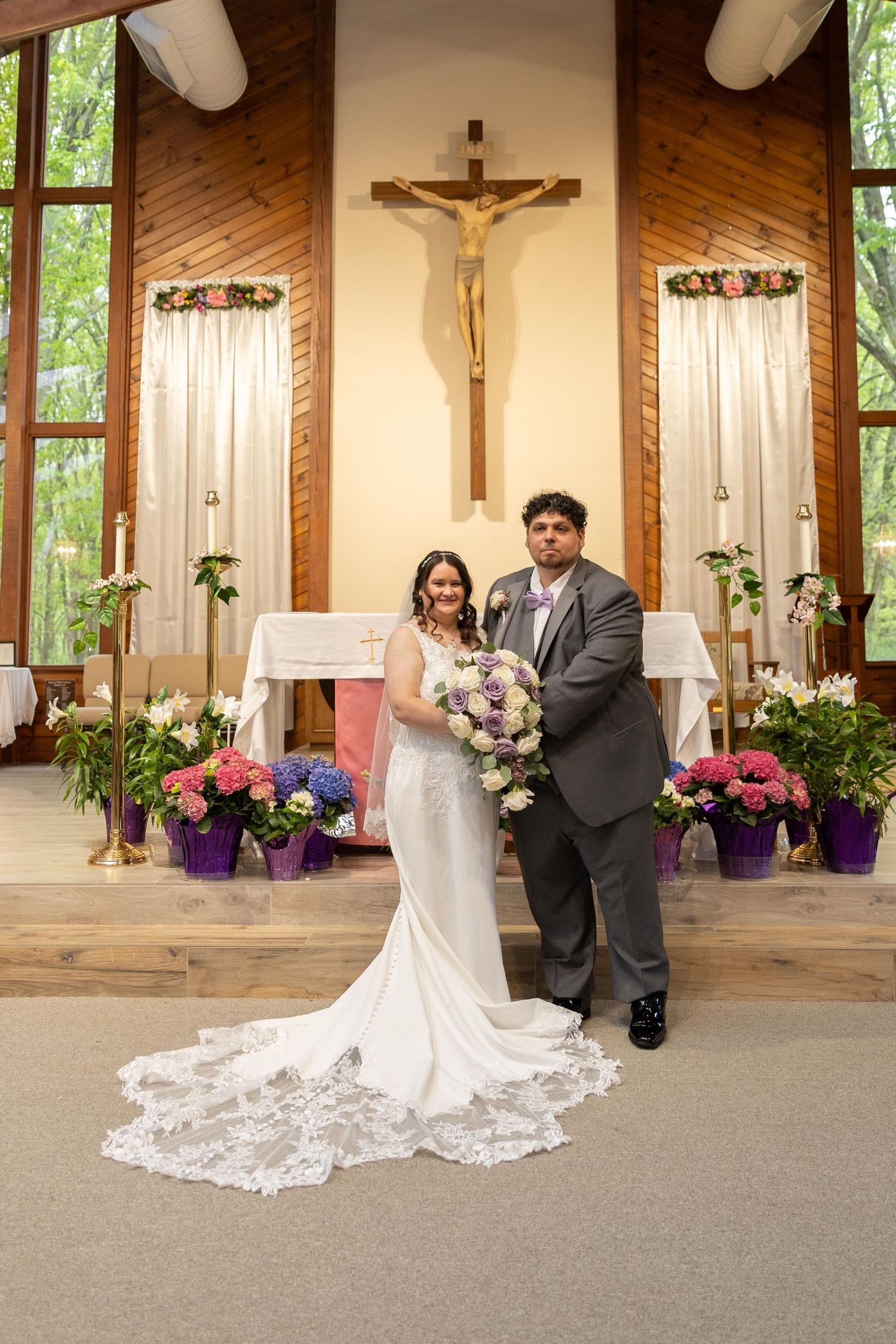 Bride and groom pose together at church altar with crucifix, floral arrangements, and lace wedding dress train displayed beautifully.