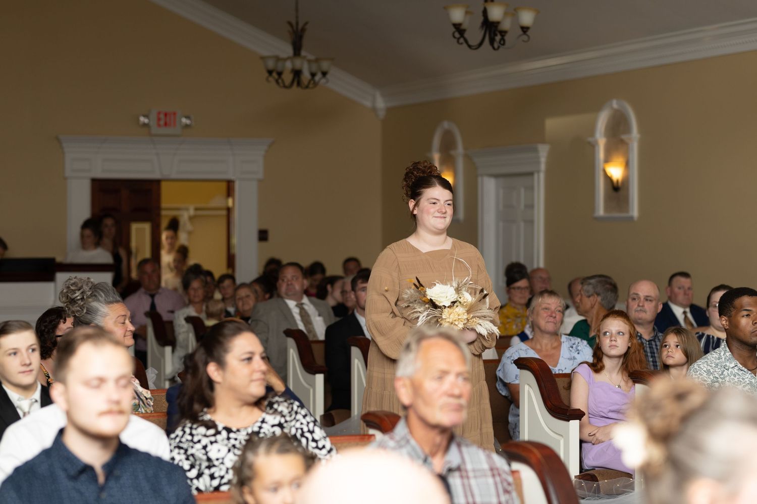 Wedding guests fill the rows of a formal ceremony space with cream colored walls and chandeliers.