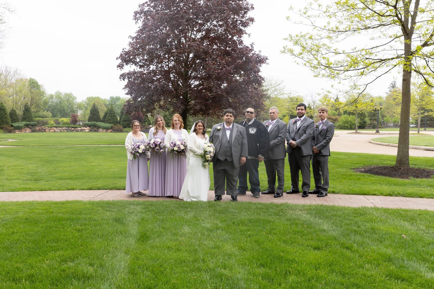 Wedding party posing outdoors on lush green lawn with bridesmaids in lavender dresses and groomsmen in gray suits.