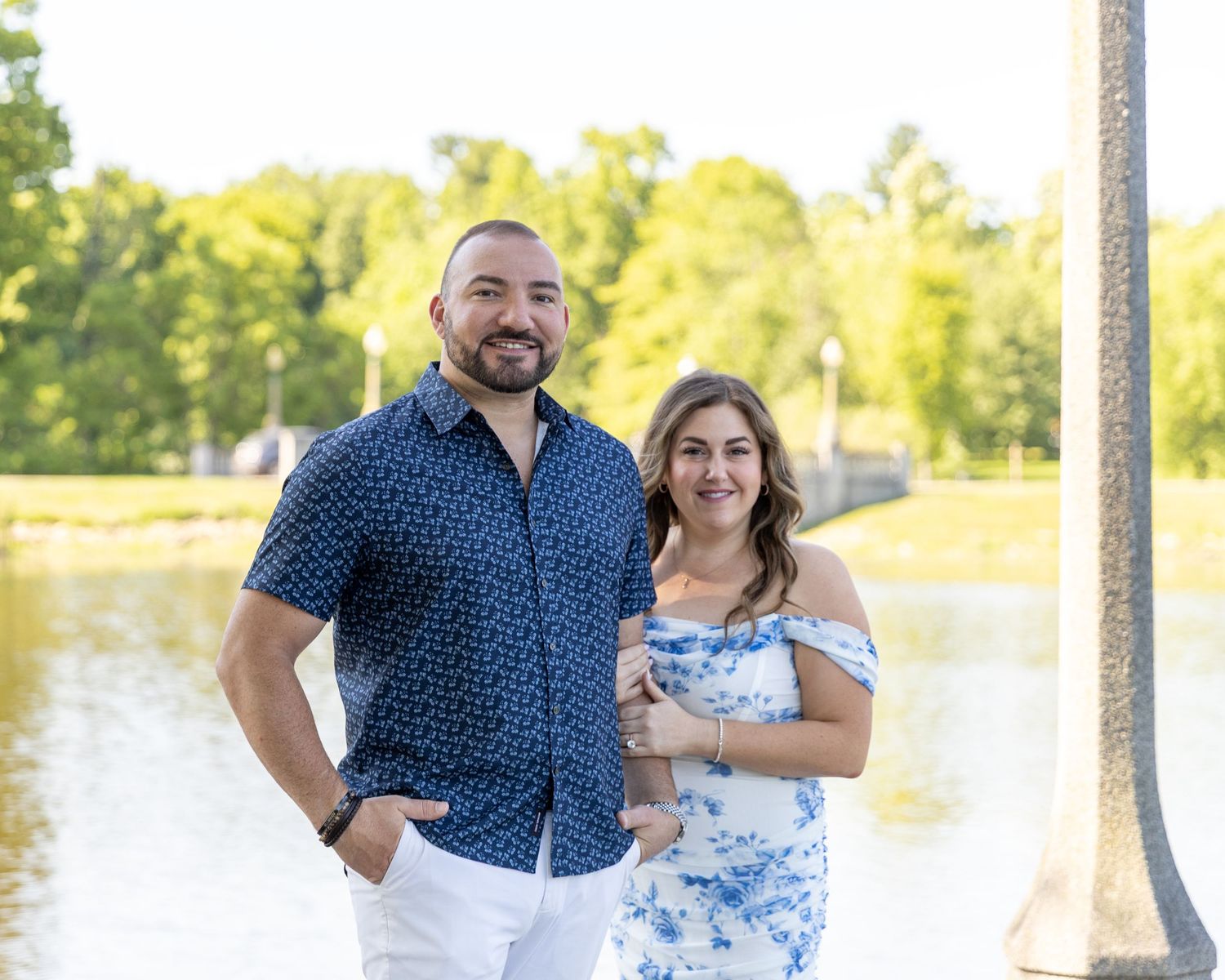 A smiling couple poses outdoors near a lake in a lush green park, wearing casual blue and white outfits.