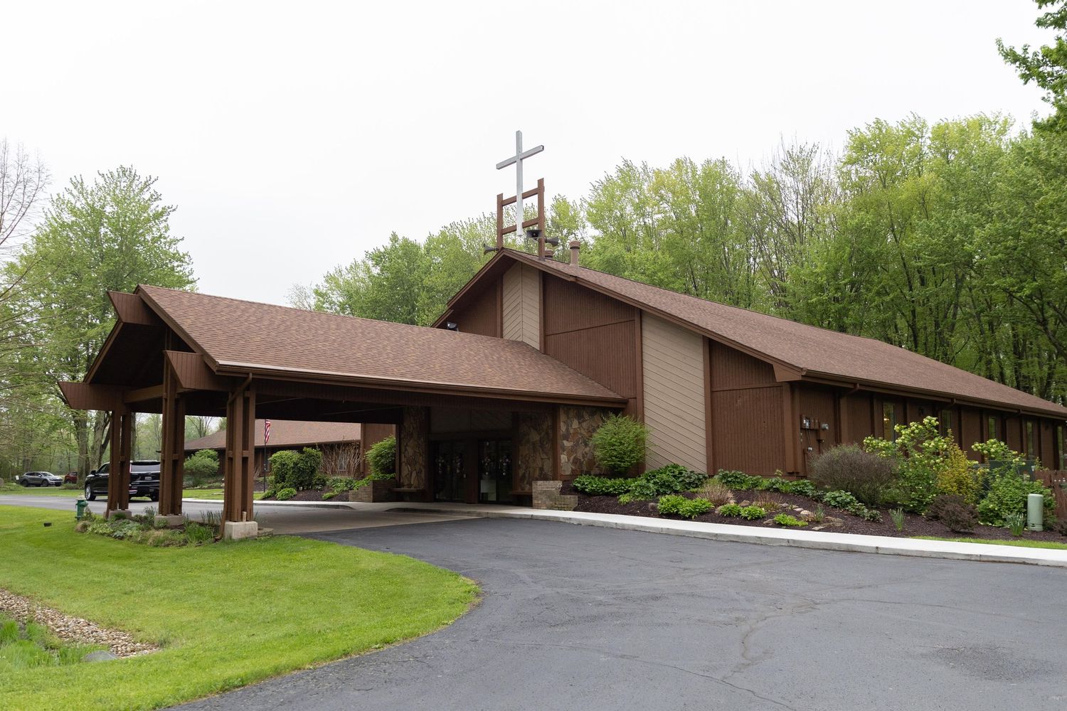 Brown wooden church with a cross on the roof, surrounded by lush green trees and a paved driveway.