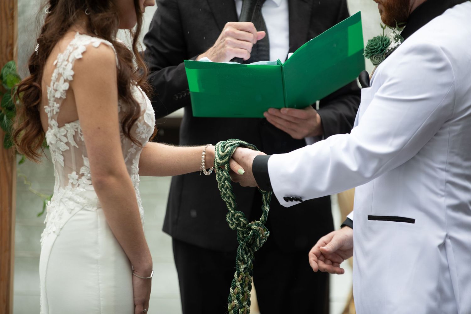 Bride and groom performing handfasting ceremony with green ribbon, officiant holding folder in background.