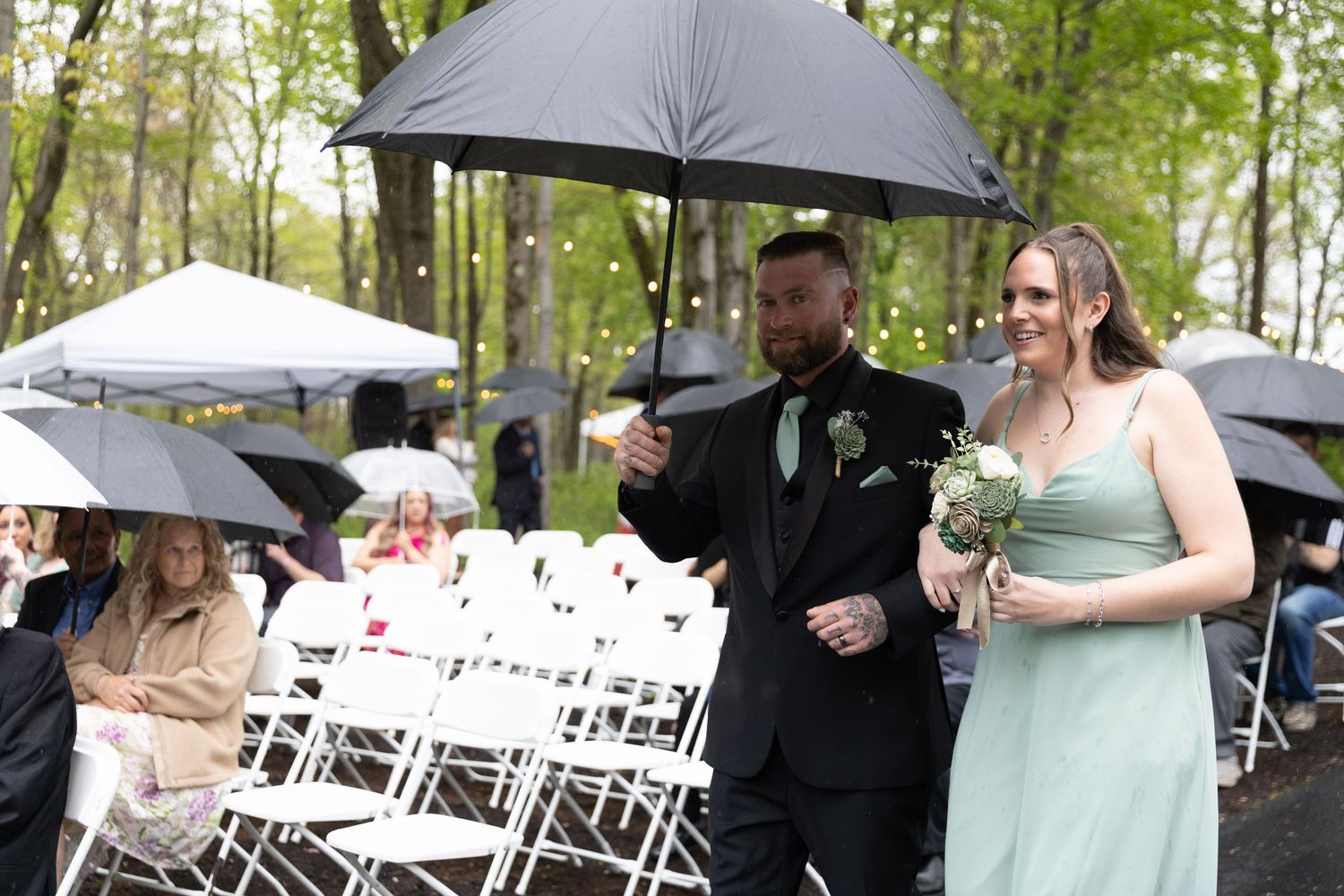 A man and woman walk down a rainy outdoor wedding aisle under a black umbrella, guests seated in white chairs.