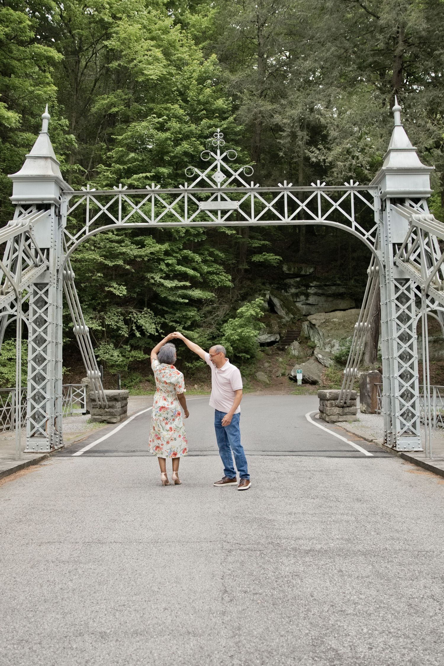 Couple dancing under a white ornate garden gate arch surrounded by green trees.
