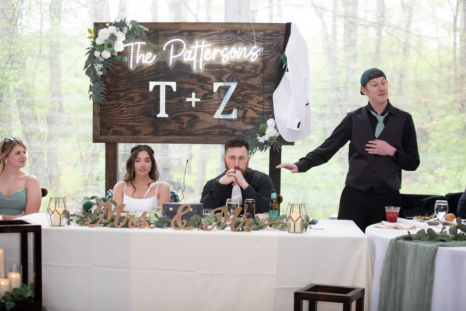 Wedding reception toast at The Pattersons T+Z head table, with guests seated beneath a rustic wooden sign.