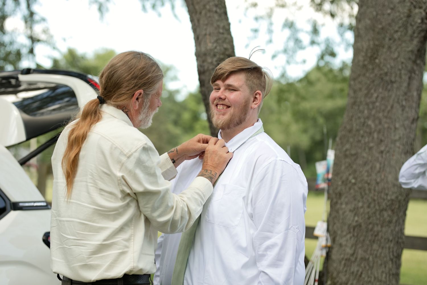 Woman adjusting a young man's tie outdoors near a tree, both dressed in white formal attire.