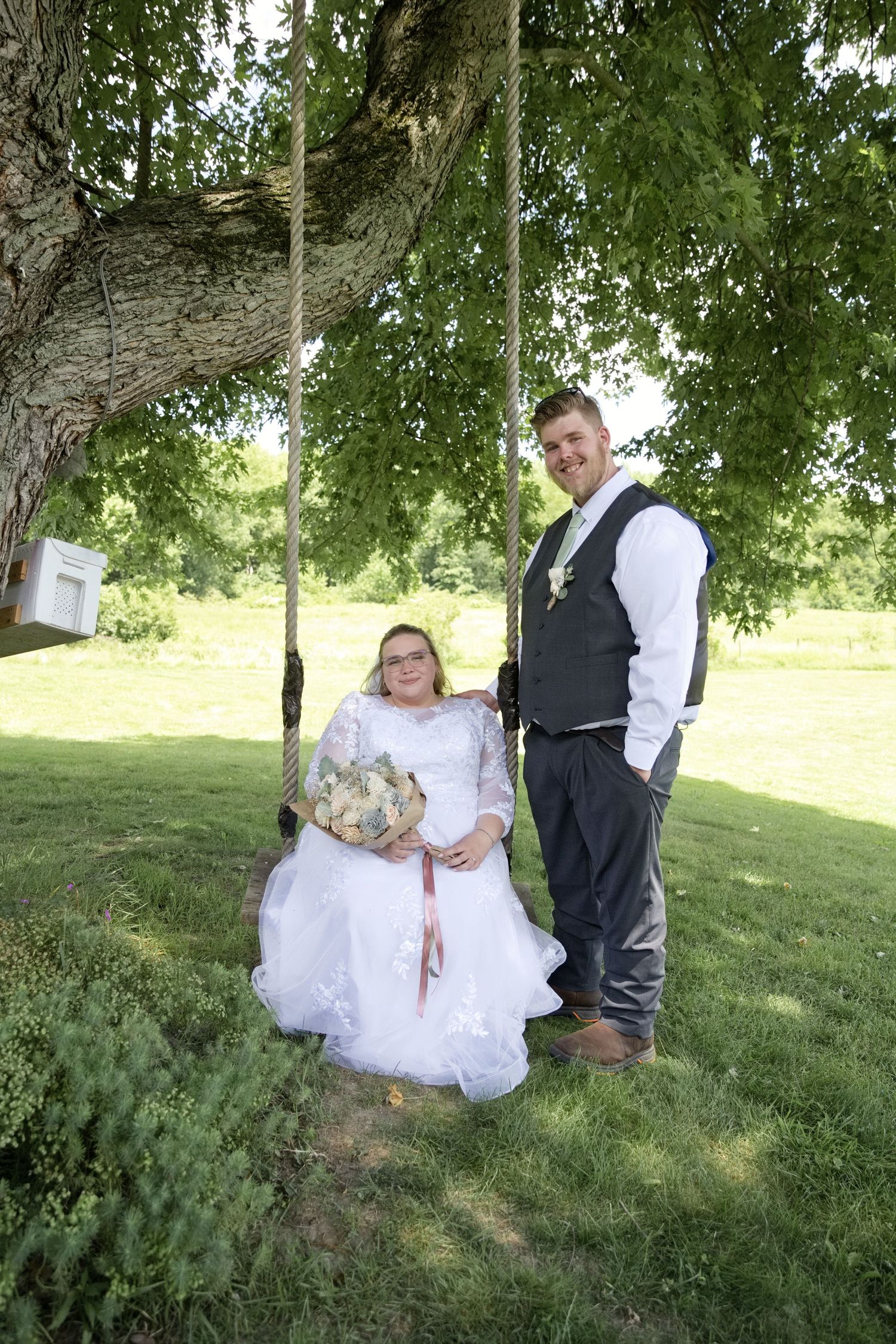 Bride sitting on tree swing holding bouquet beside groom in vest, posing outdoors on green lawn for wedding portrait.