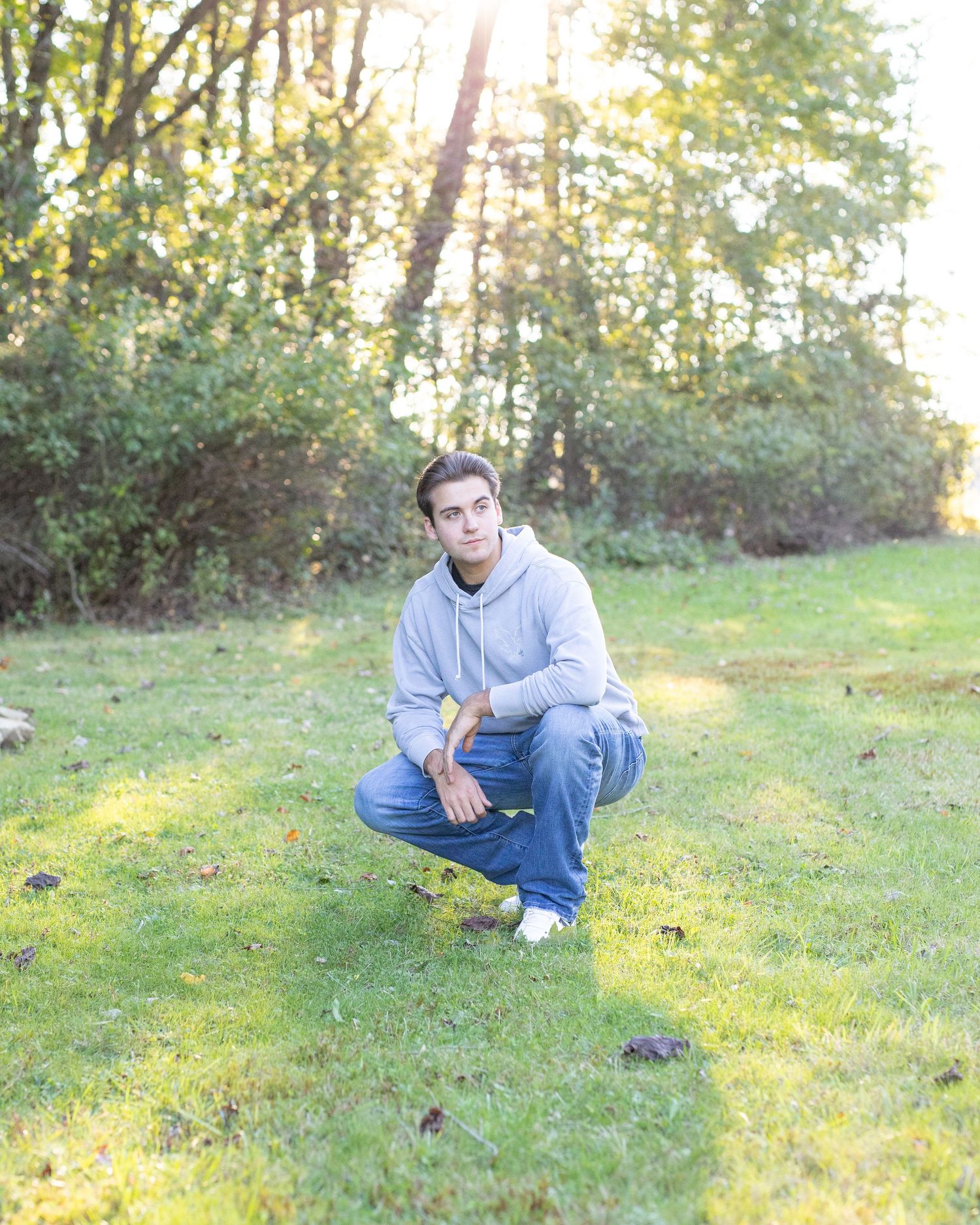 Young man in a light gray hoodie and jeans on a sunlit grassy field, with a forested background and sunlight streaming through the trees