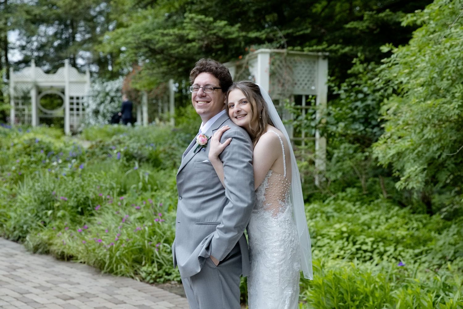 Bride and groom smiling together in a lush garden, bride wearing a white veil and gown, groom in a gray suit.