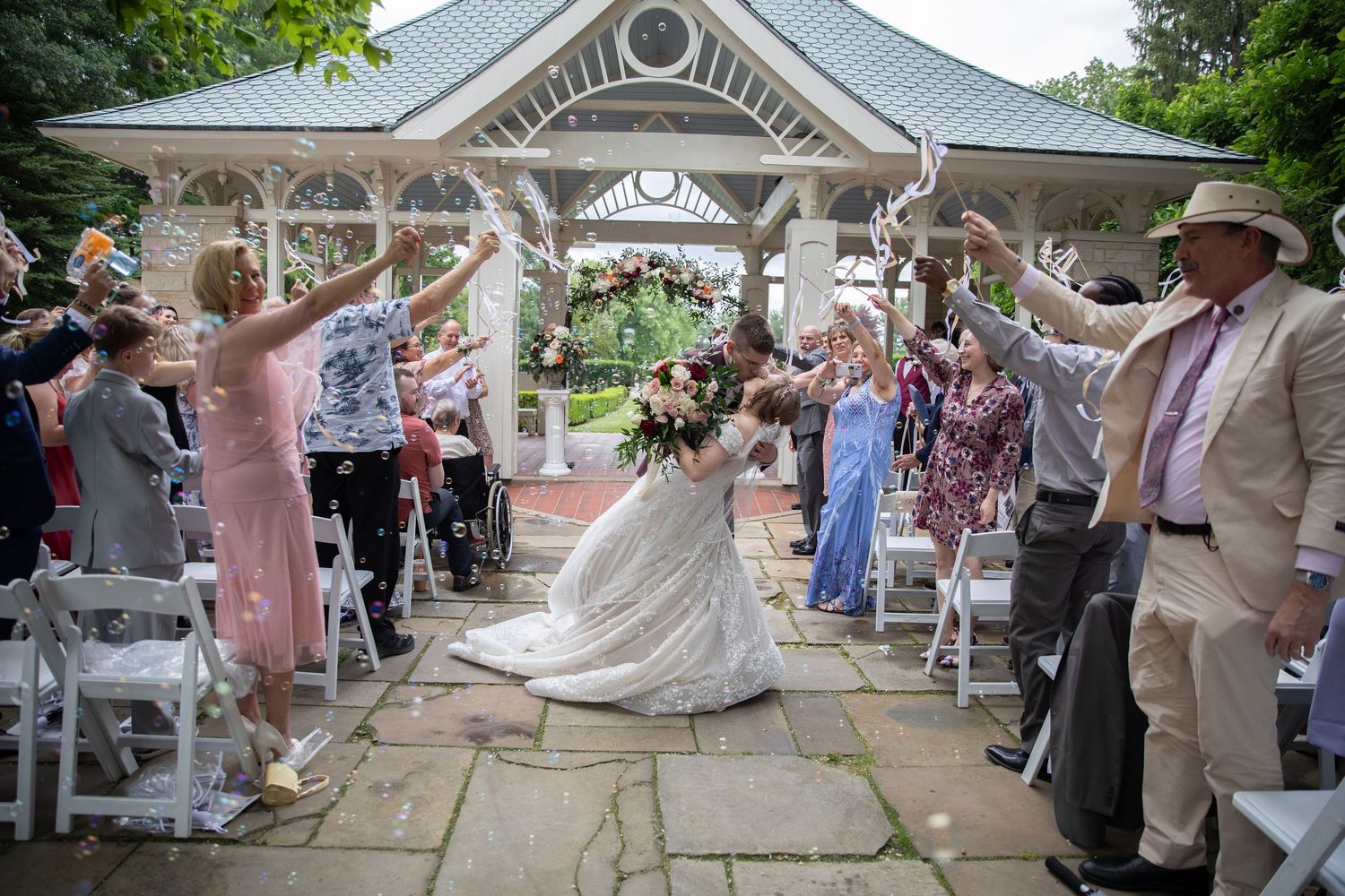 Bride walks down aisle under raised hands at outdoor gazebo wedding ceremony with guests celebrating.