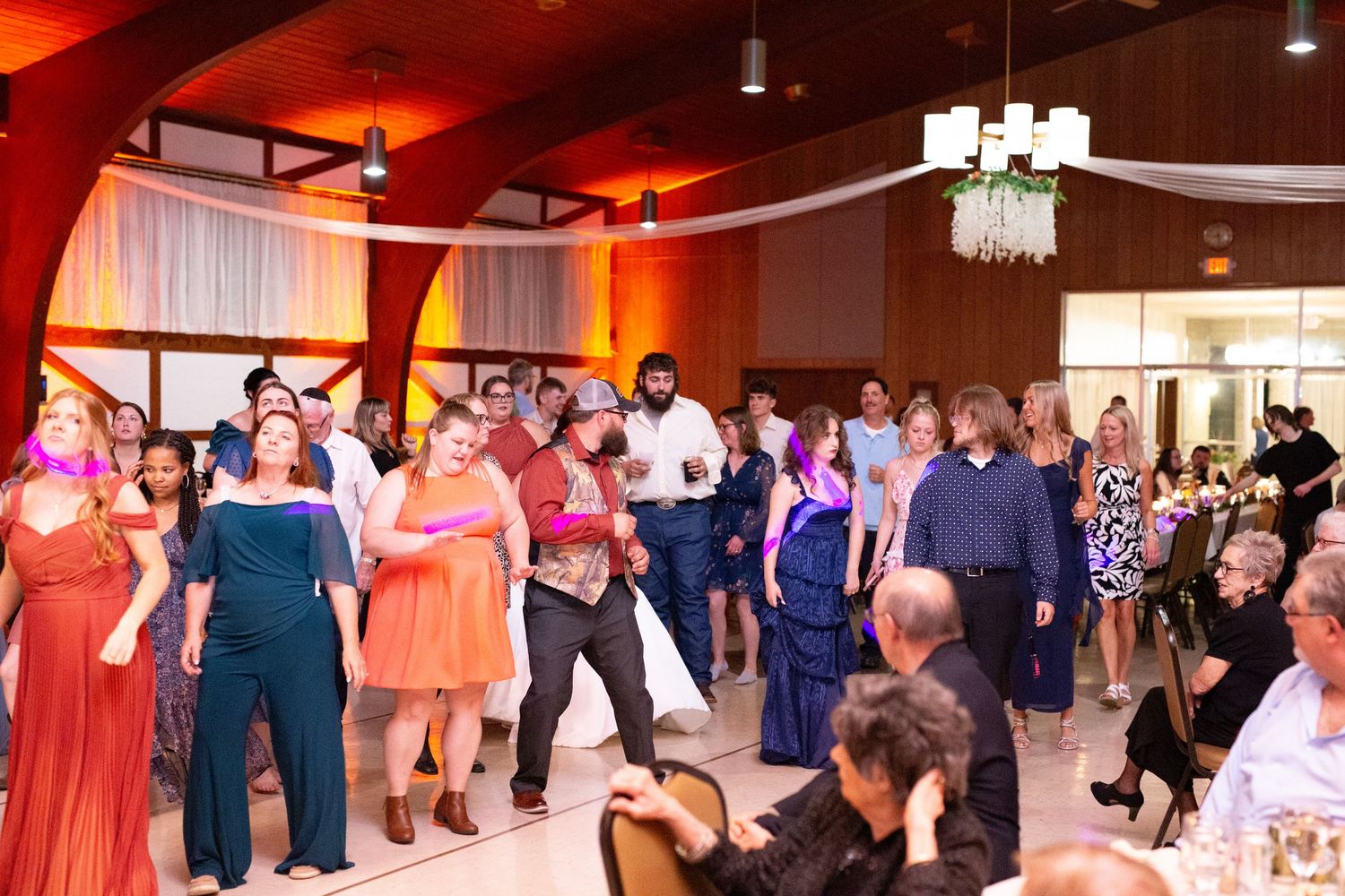 Large group of wedding guests dancing together on illuminated reception dance floor.