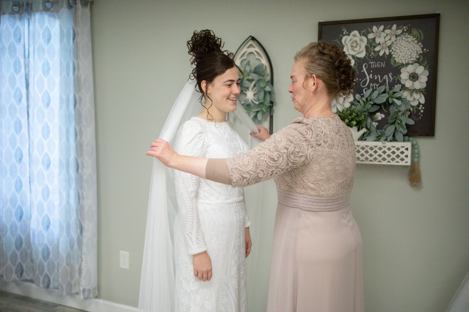 A touching pre-wedding moment between two people in a room with lace curtains and floral artwork.