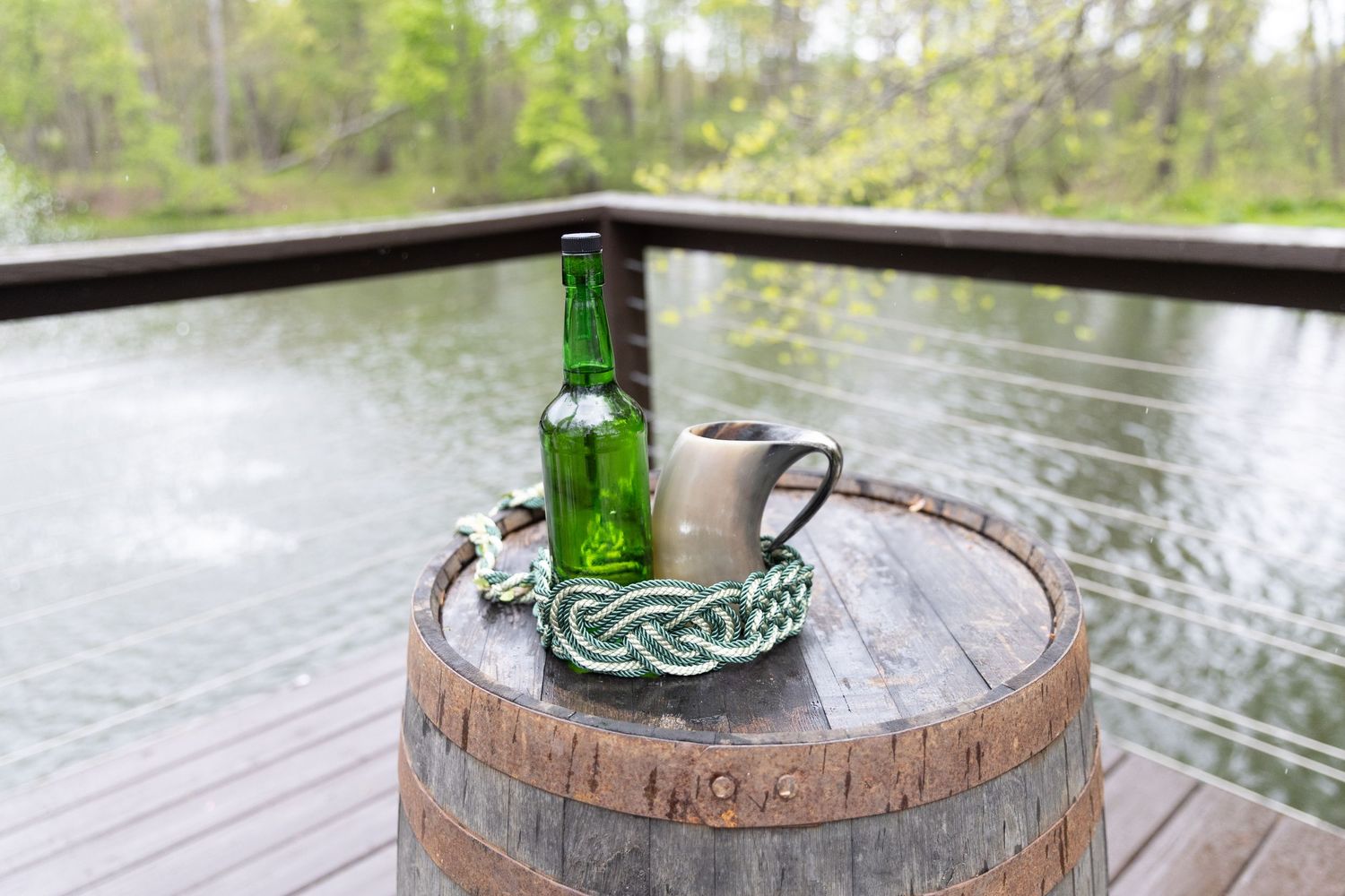 Green beer bottle and metal mug on rustic wooden barrel on deck overlooking serene forest pond.