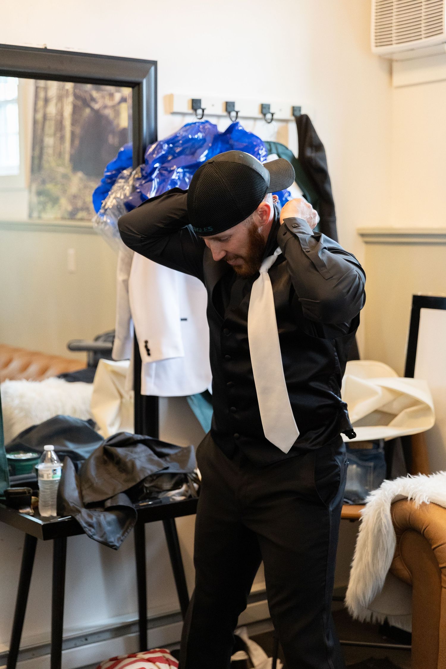 Man in black shirt and tie adjusting his collar while getting dressed in a room with suits hanging in background.