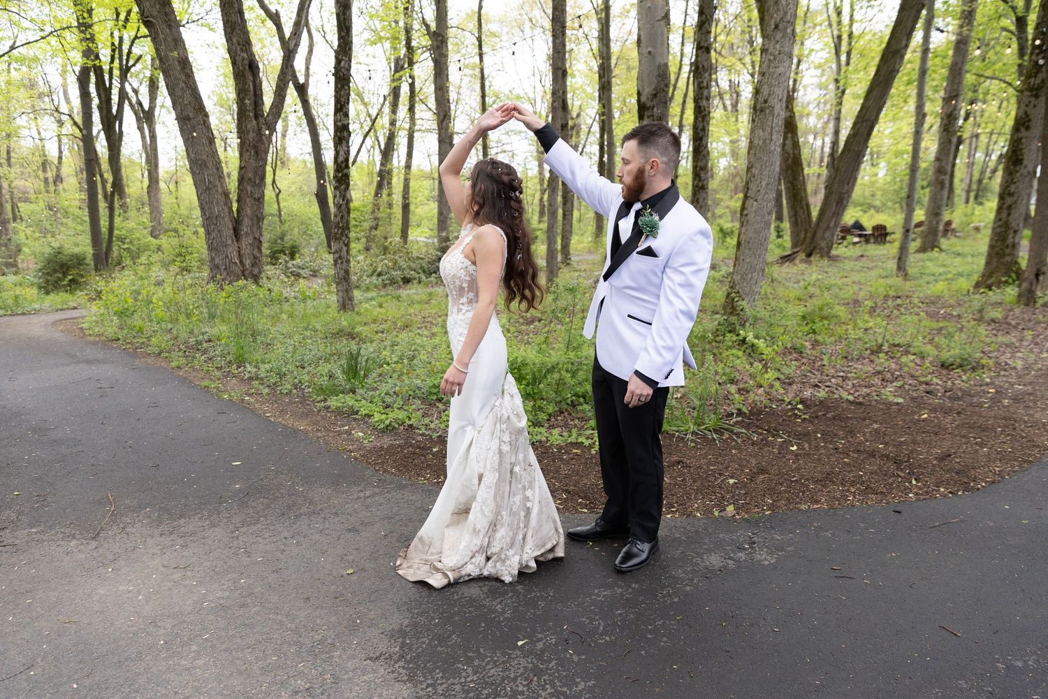 Bride and groom dancing outdoors on a path surrounded by lush green trees in a romantic wedding photo.