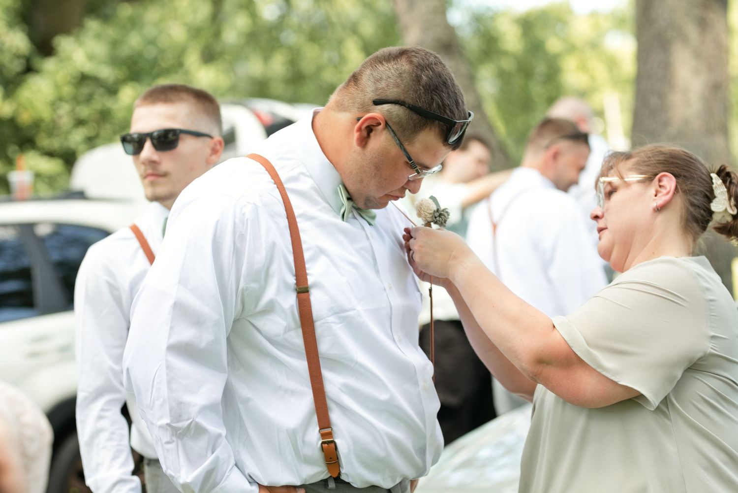 Woman pinning a boutonniere on a groomsman wearing brown suspenders outdoors before a wedding.