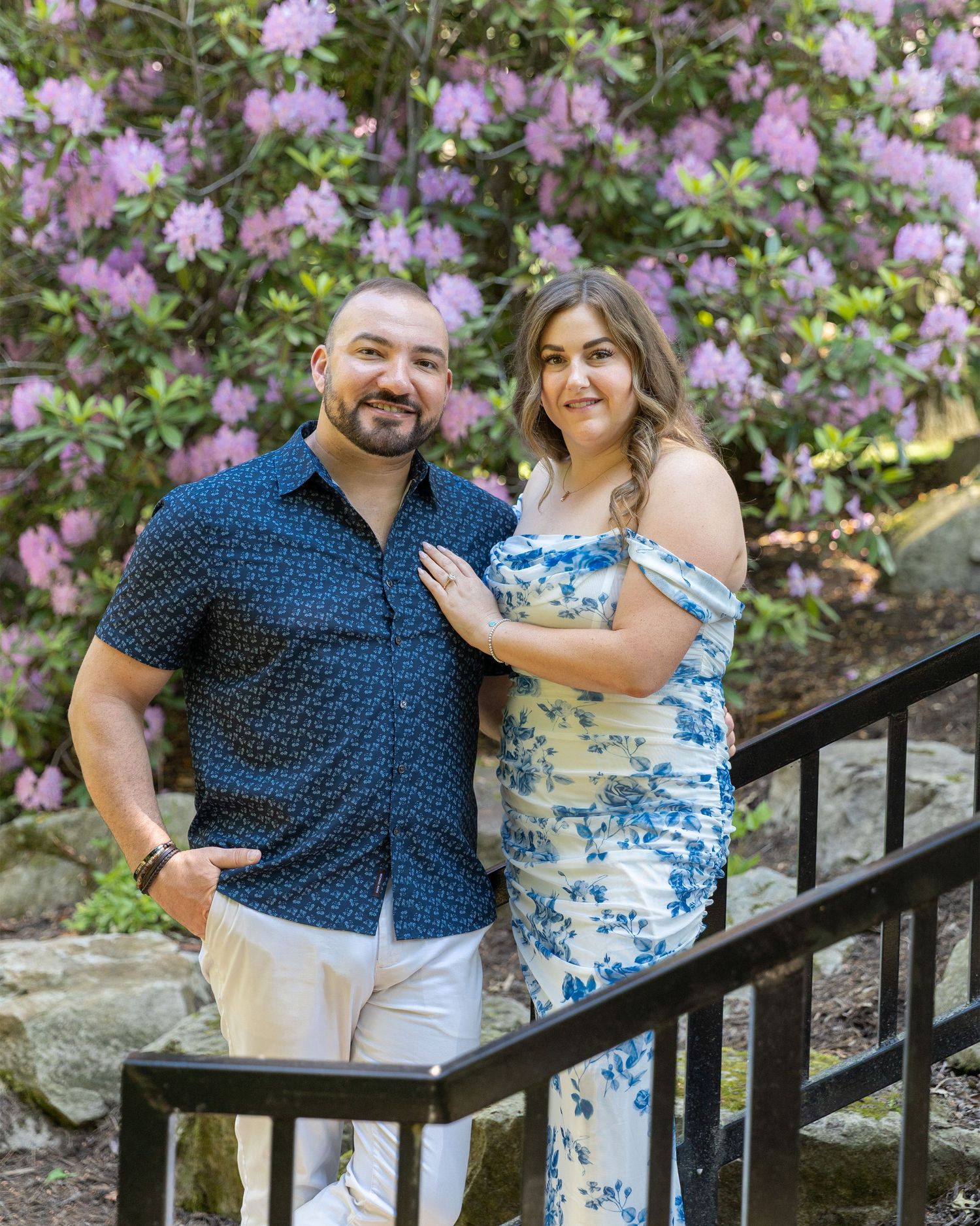 Couple posing together in front of blooming purple rhododendrons, woman in blue floral dress, man in navy shirt.