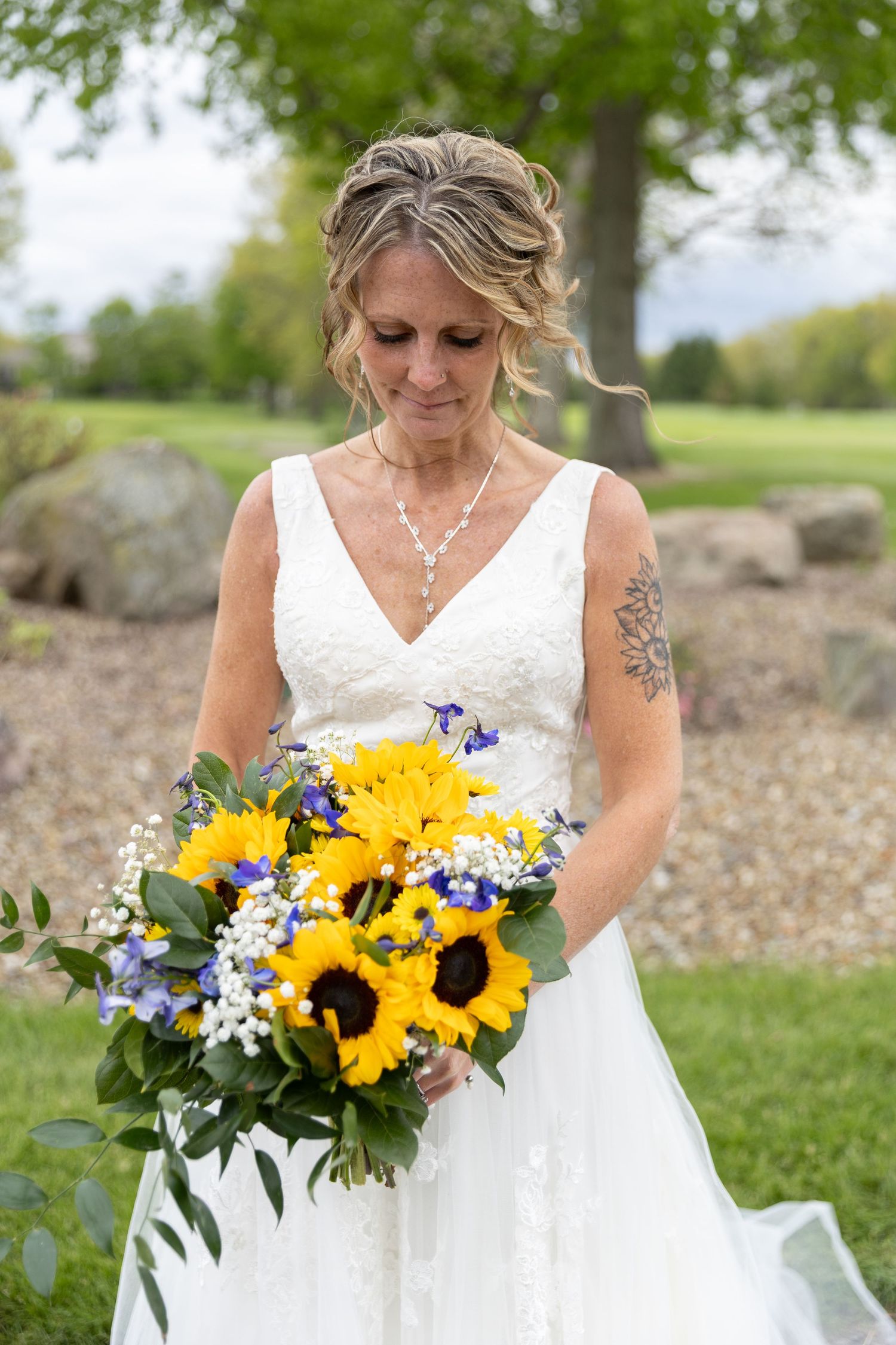 Bride in white lace gown holds vibrant sunflower bouquet with blue accents, smiling downward outdoors.