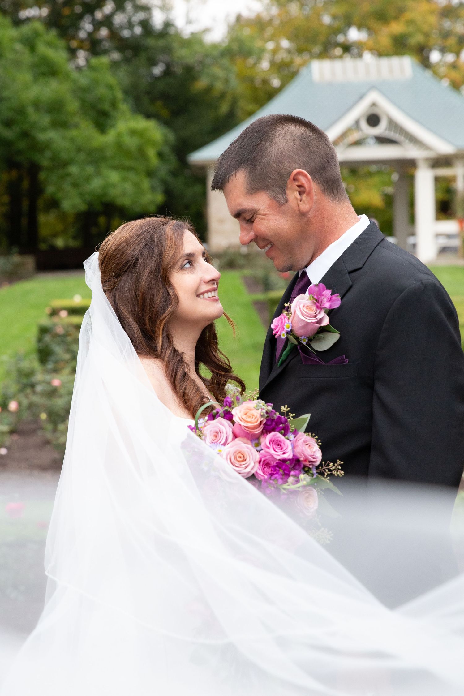 Romantic wedding couple poses in a garden setting with pink roses and flowing veil.