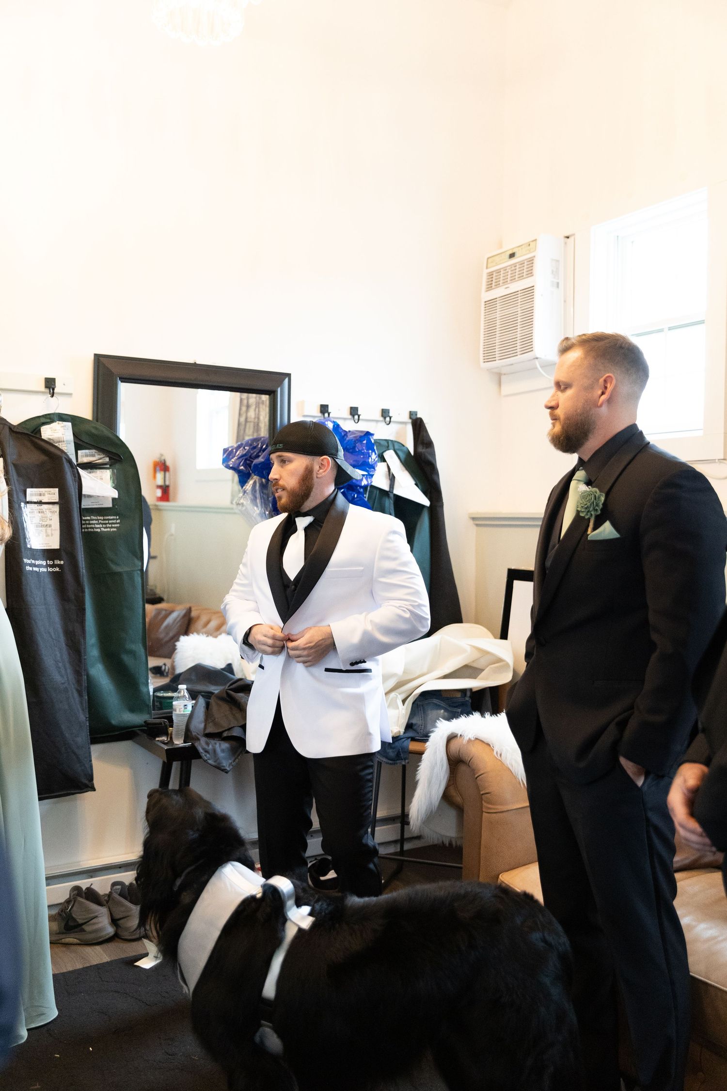 Two grooms in formal attire prepare for their wedding day, with a large black dog nearby in a dressing room.