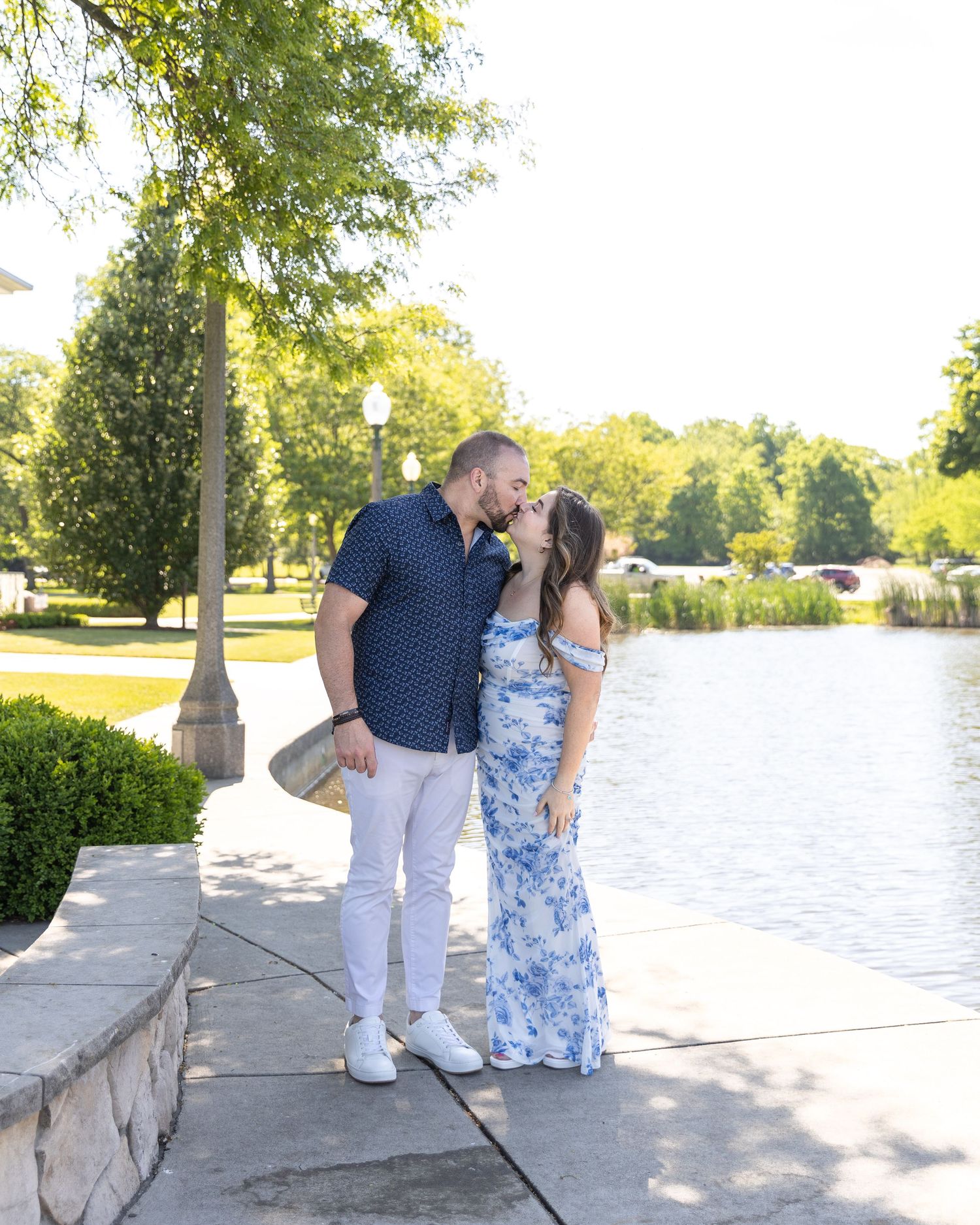 Couple sharing a kiss by a serene lake in a sunny park, dressed in coordinating outfits.