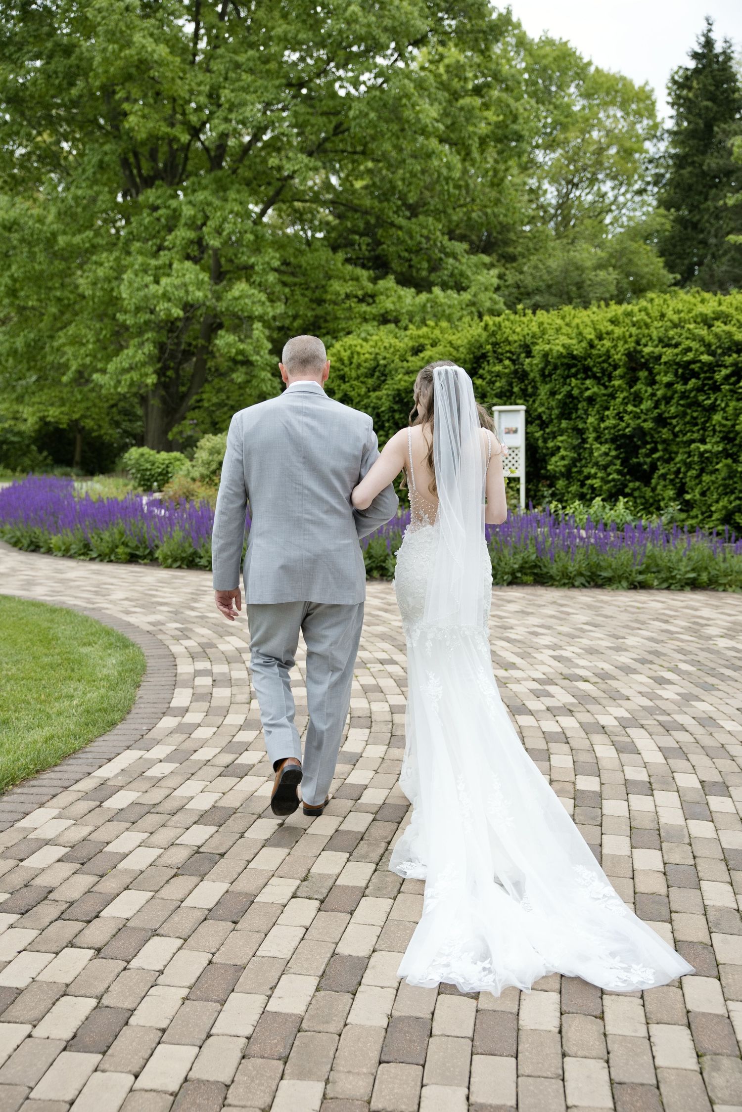 Bride and groom walking hand in hand on a brick path through a lush garden, bride wearing a white gown with a long train.