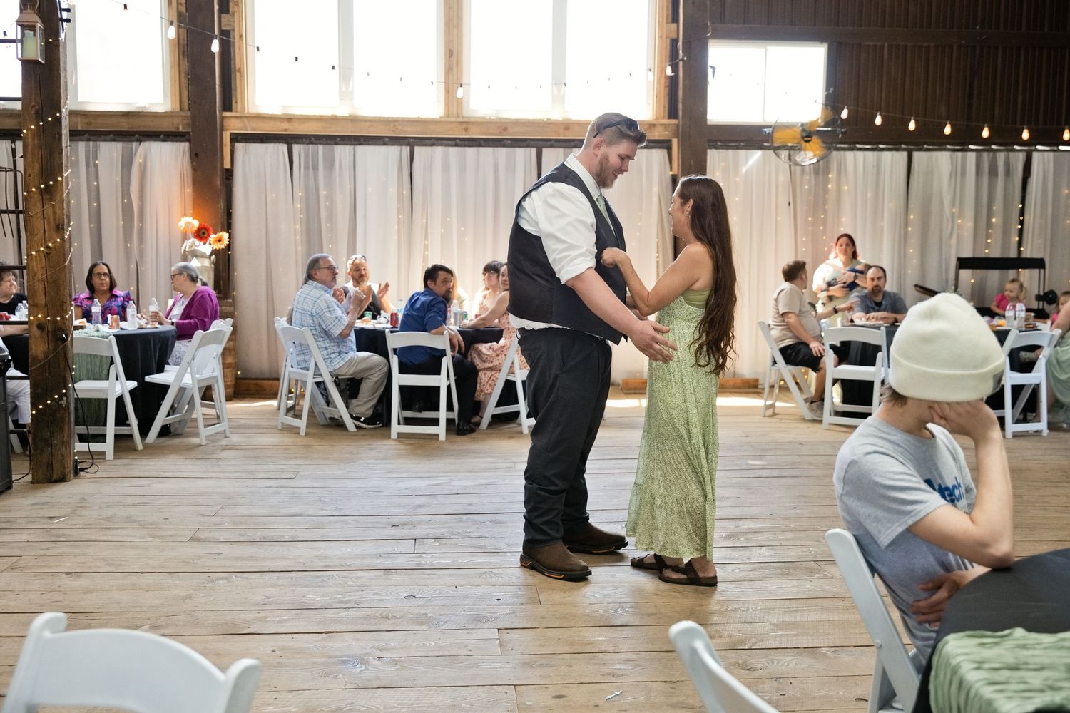 Couple shares first dance in rustic barn wedding venue with guests seated at white chairs in background.