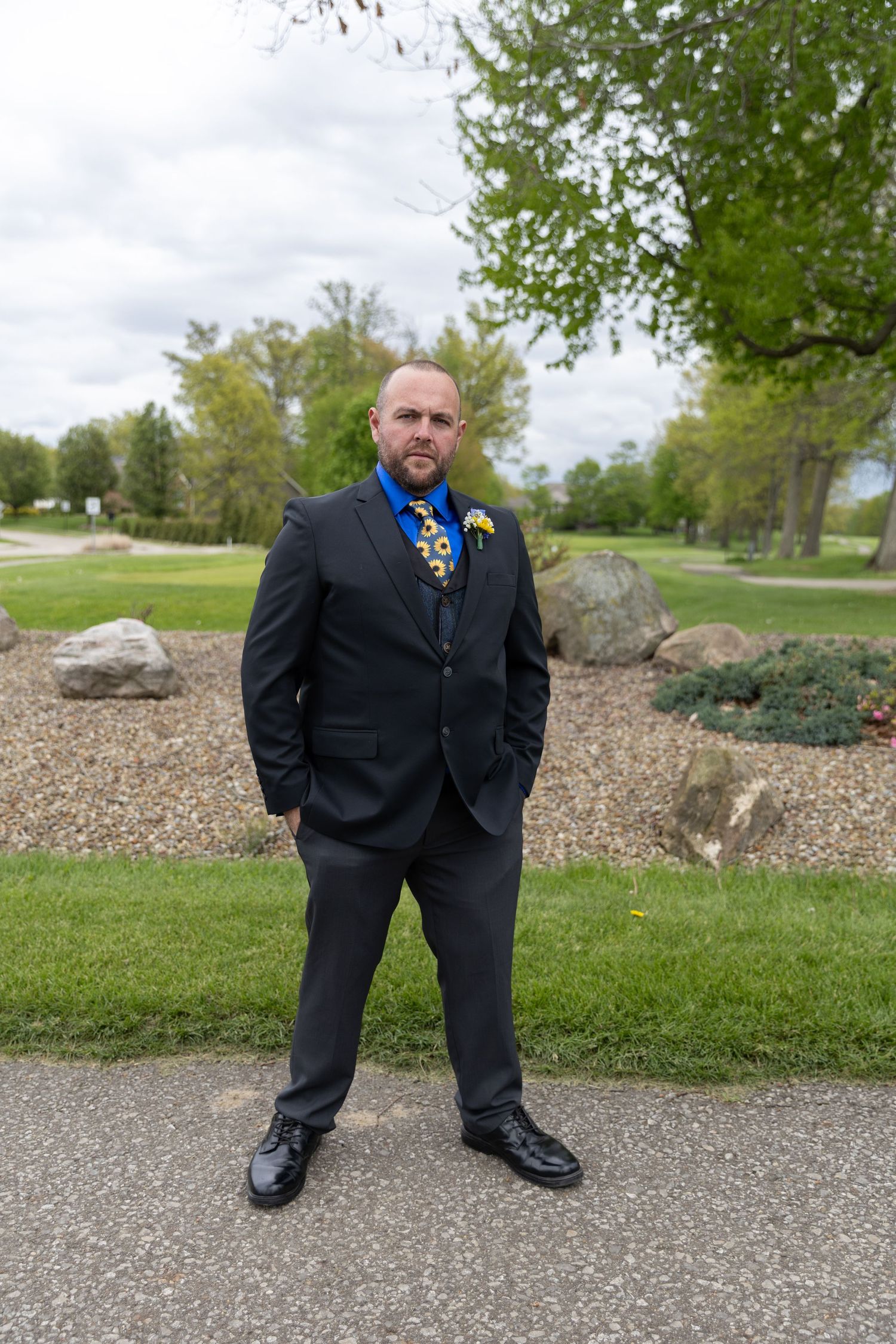 Bald groom in dark suit with blue tie and boutonniere stands confidently outdoors in a park setting.