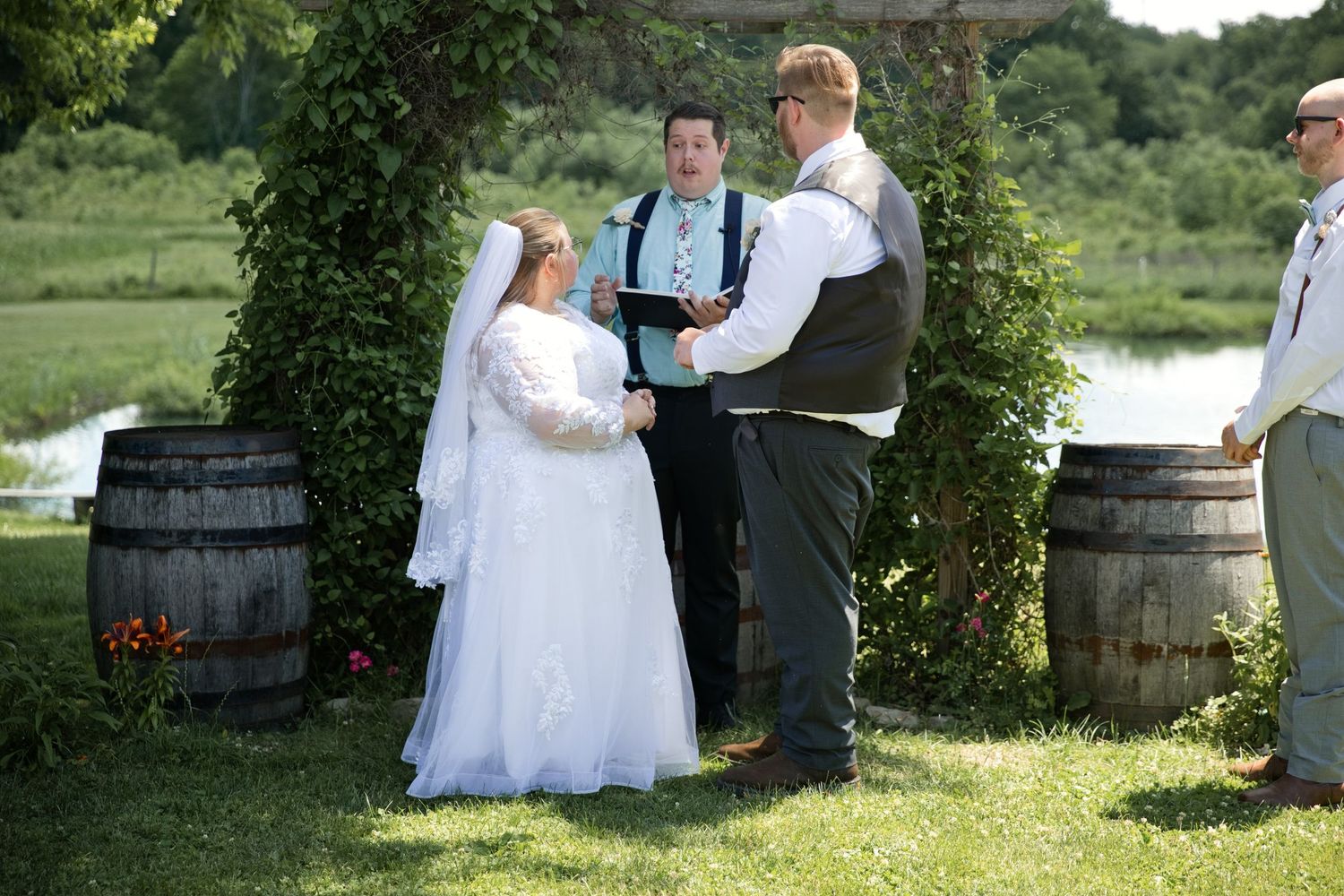 Bride and groom hold hands during outdoor wedding ceremony between rustic barrels with officiant and lush greenery.