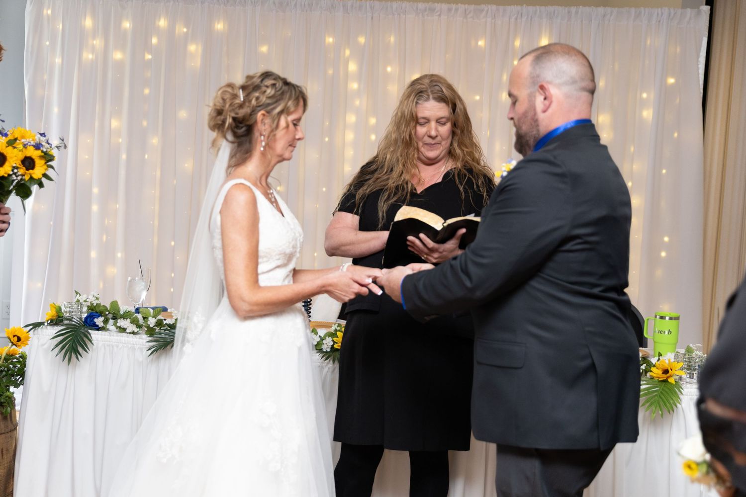 Bride and groom exchanging vows at altar with officiant holding Bible, decorated with sunflowers and fairy lights.