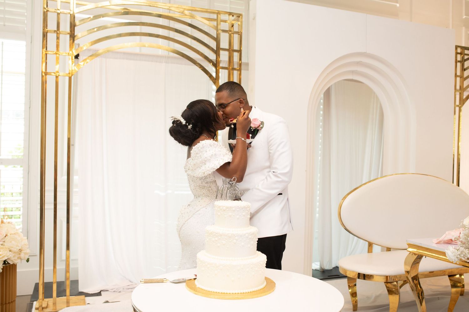 Elegant couple sharing a romantic moment next to their wedding cake in a luxurious white and gold venue.