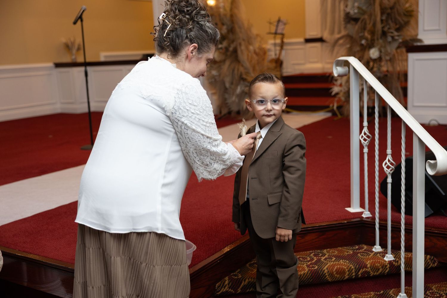 A person in white helps a young child in a brown suit with formal attire on red carpeted stairs.