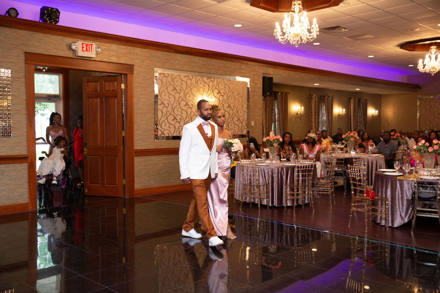 Wedding guests dancing in an elegant reception venue with purple accent lighting and chandeliers.