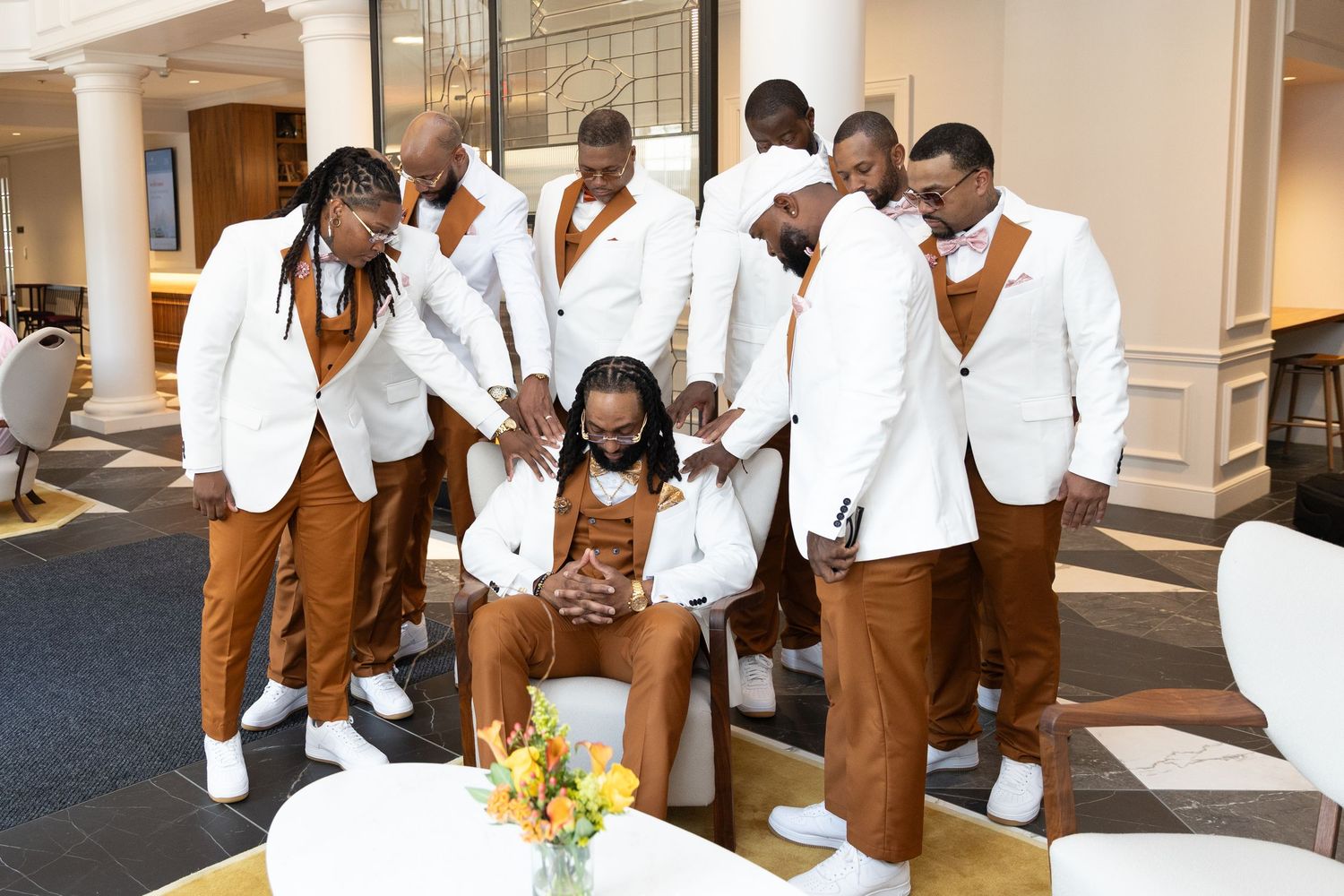 Wedding party in white jackets and brown pants gathered around seated person in formal attire.