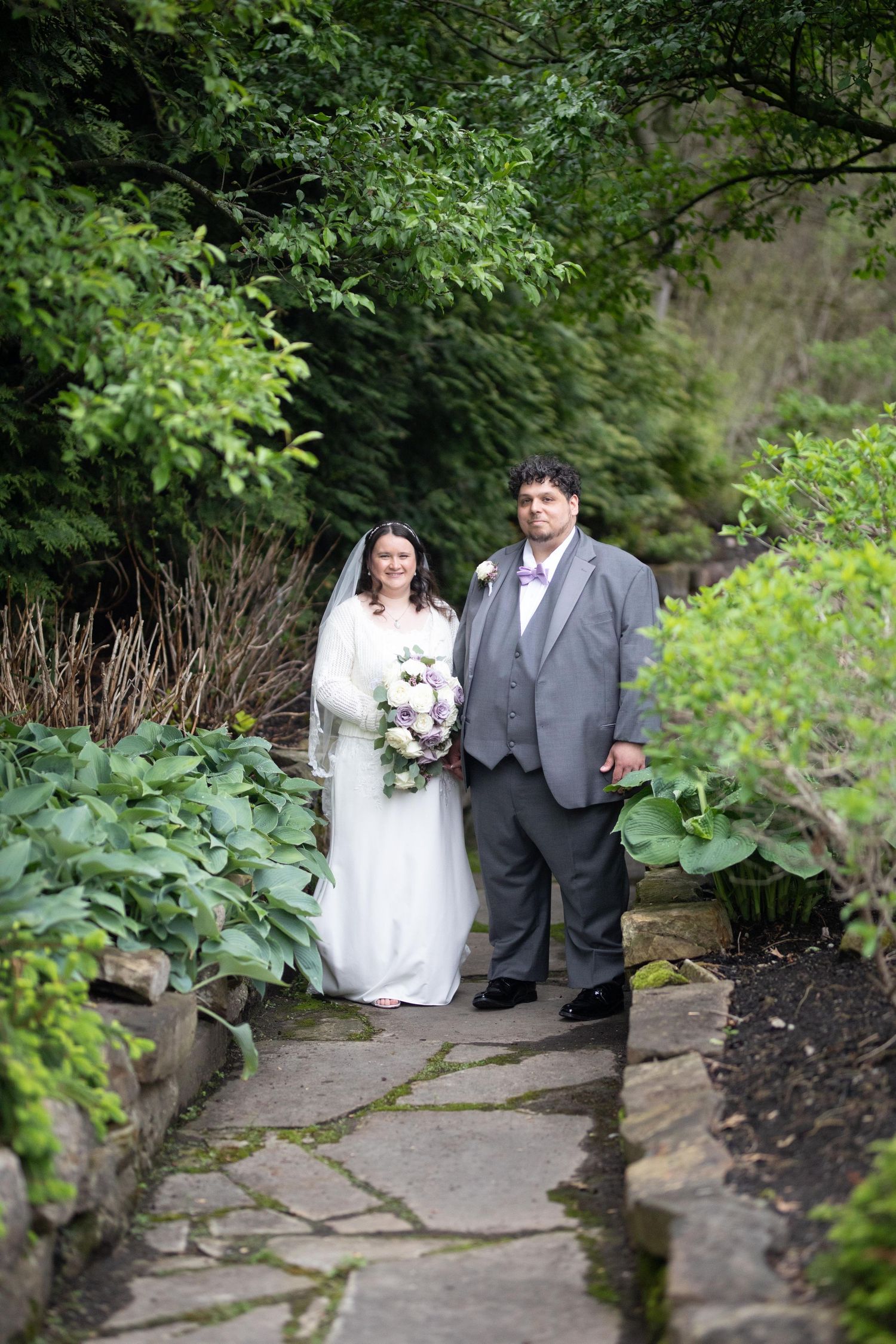 Bride and groom posing on a stone garden path surrounded by lush green foliage on their wedding day.