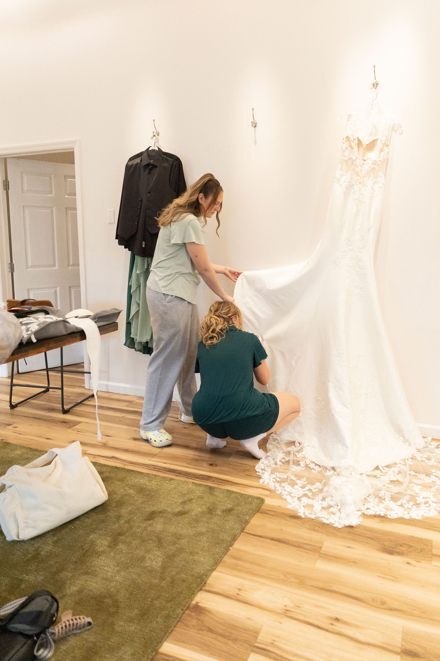 Two women adjusting a white lace wedding dress hanging in a bright bridal boutique with hardwood floors.