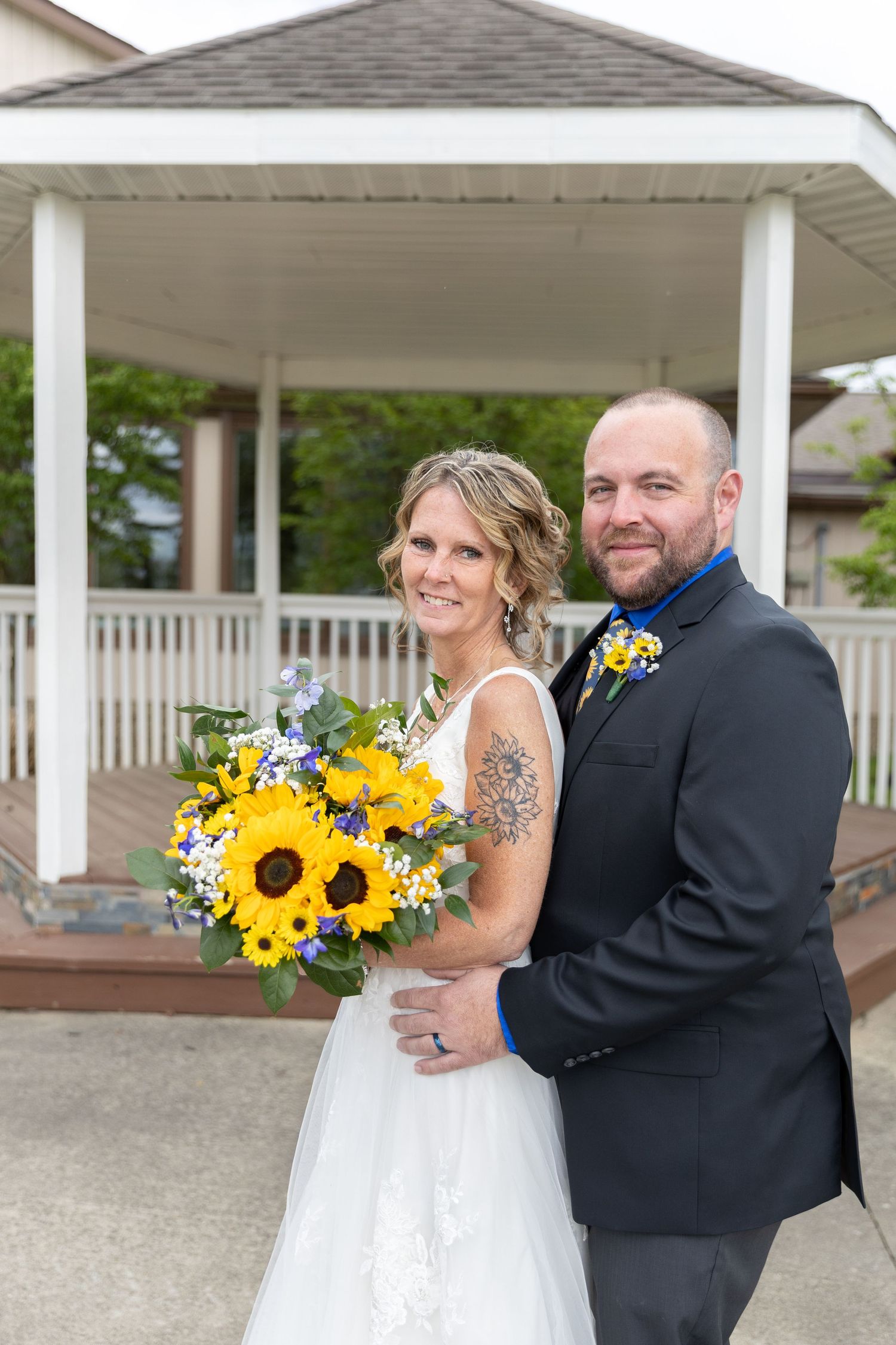 Bride and groom pose with sunflower bouquet in front of white gazebo on their wedding day.