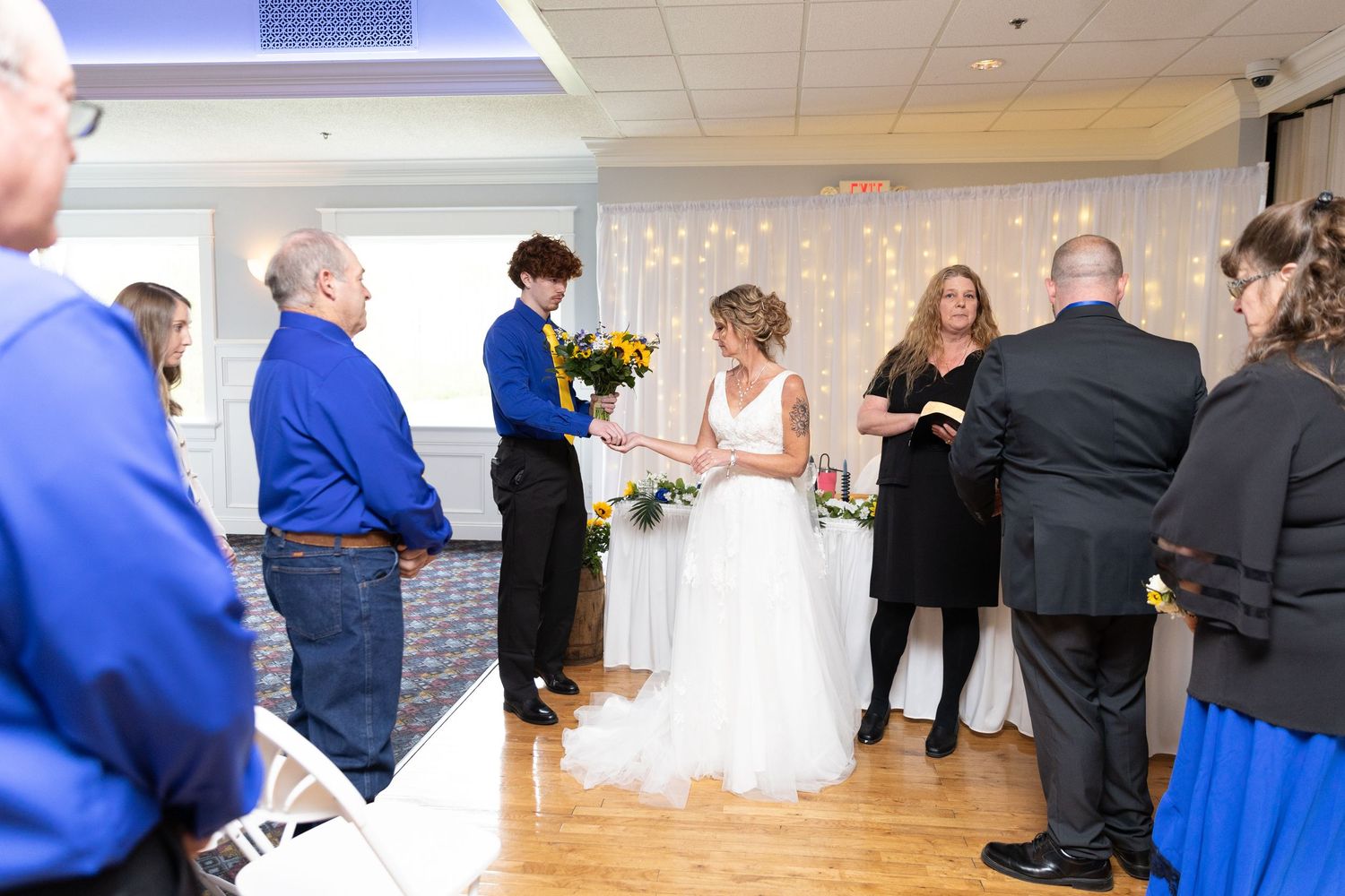 Bride in white gown during indoor wedding ceremony with guests in blue and black attire, decorated with string lights.