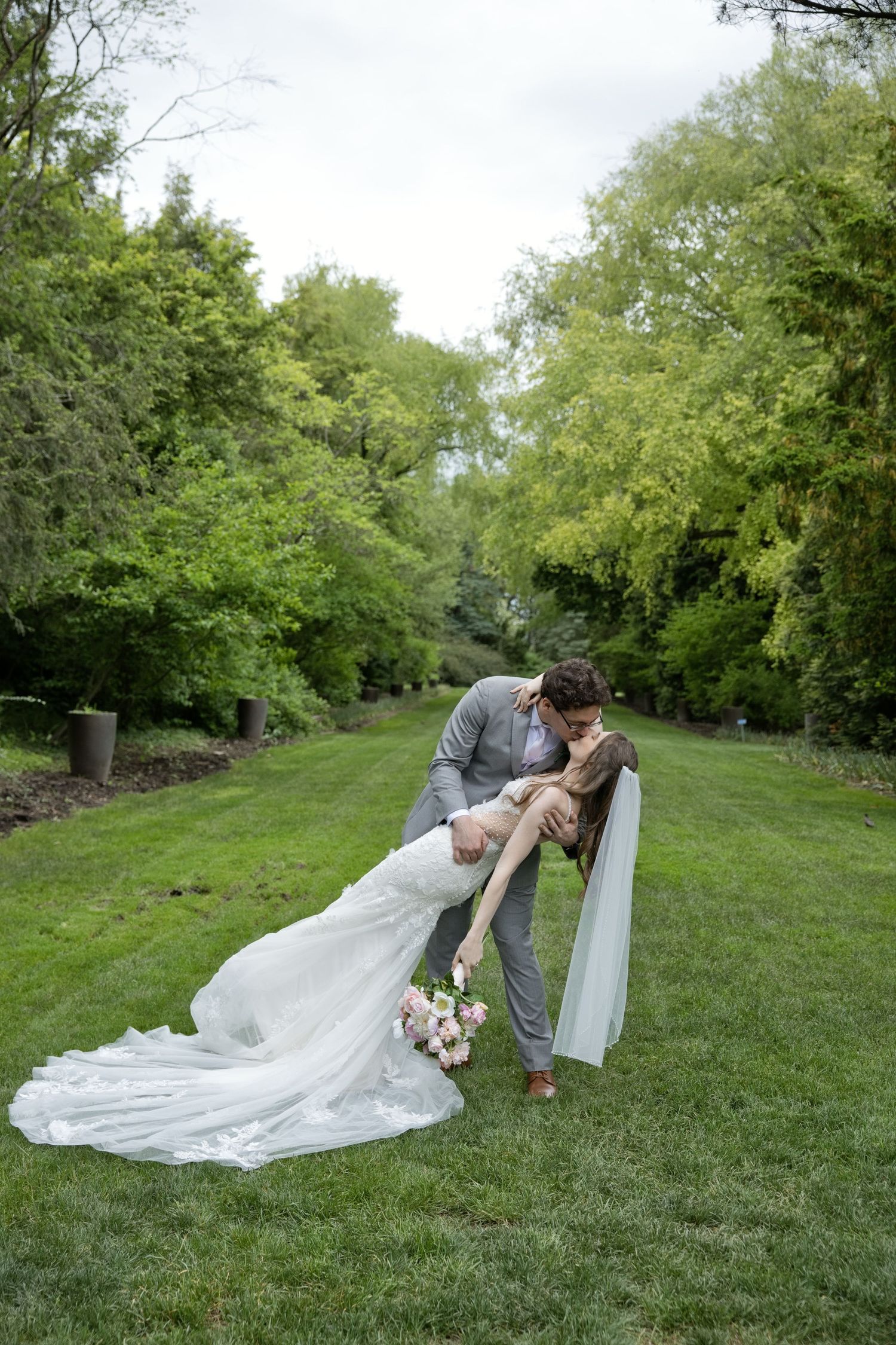 Groom dips bride in white gown for romantic kiss in lush garden setting.